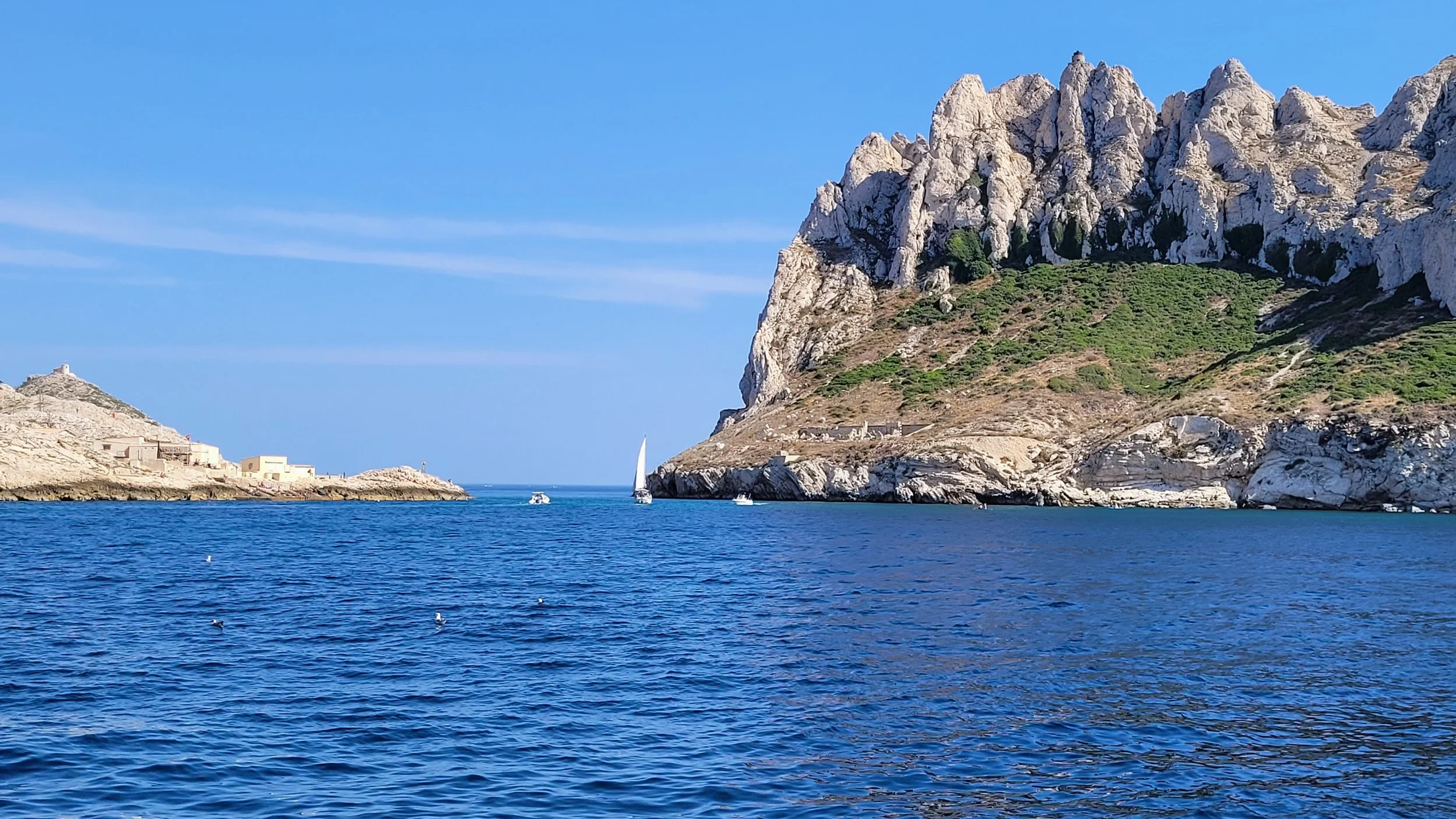 A scenic coastal landscape with a large rocky hill or mountain on the right side and smaller rocks or structures on the left. The blue ocean water in the foreground has a few small boats and is under a clear blue sky.