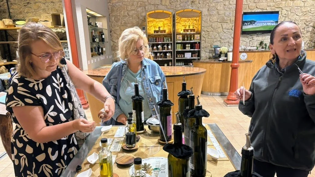 Group of three women participating in a tasting event with bottles of olive oil and balsamic vinegar on a table in a store or tasting room.