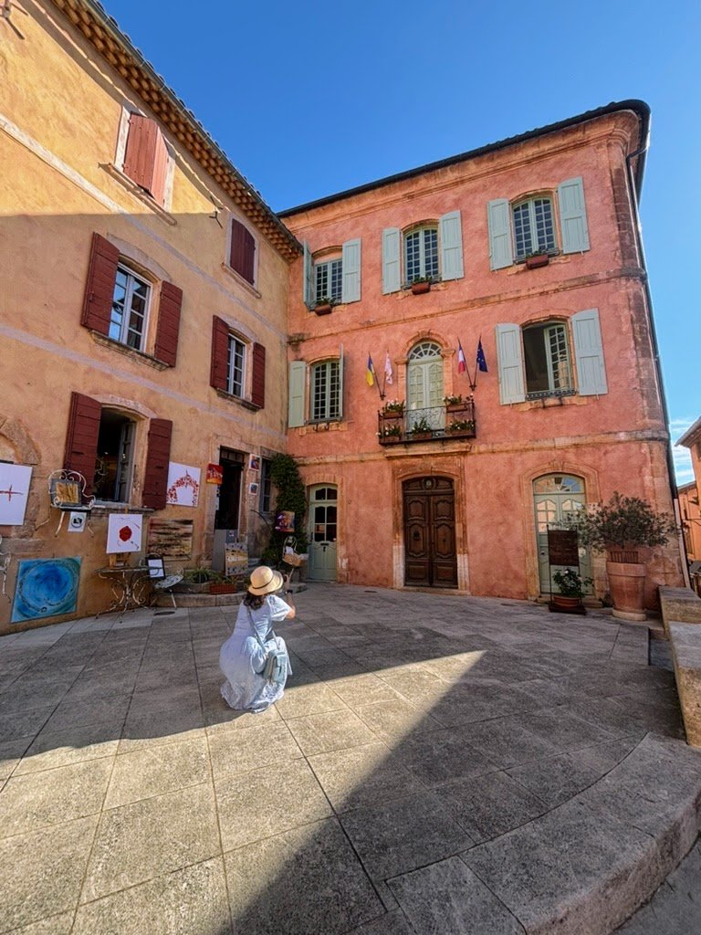 A person in a white dress and straw hat crouching on a cobblestone plaza in front of colorful old buildings with shutters, flags, and artwork displayed outside on a sunny day in Roussillon, Luberon. France