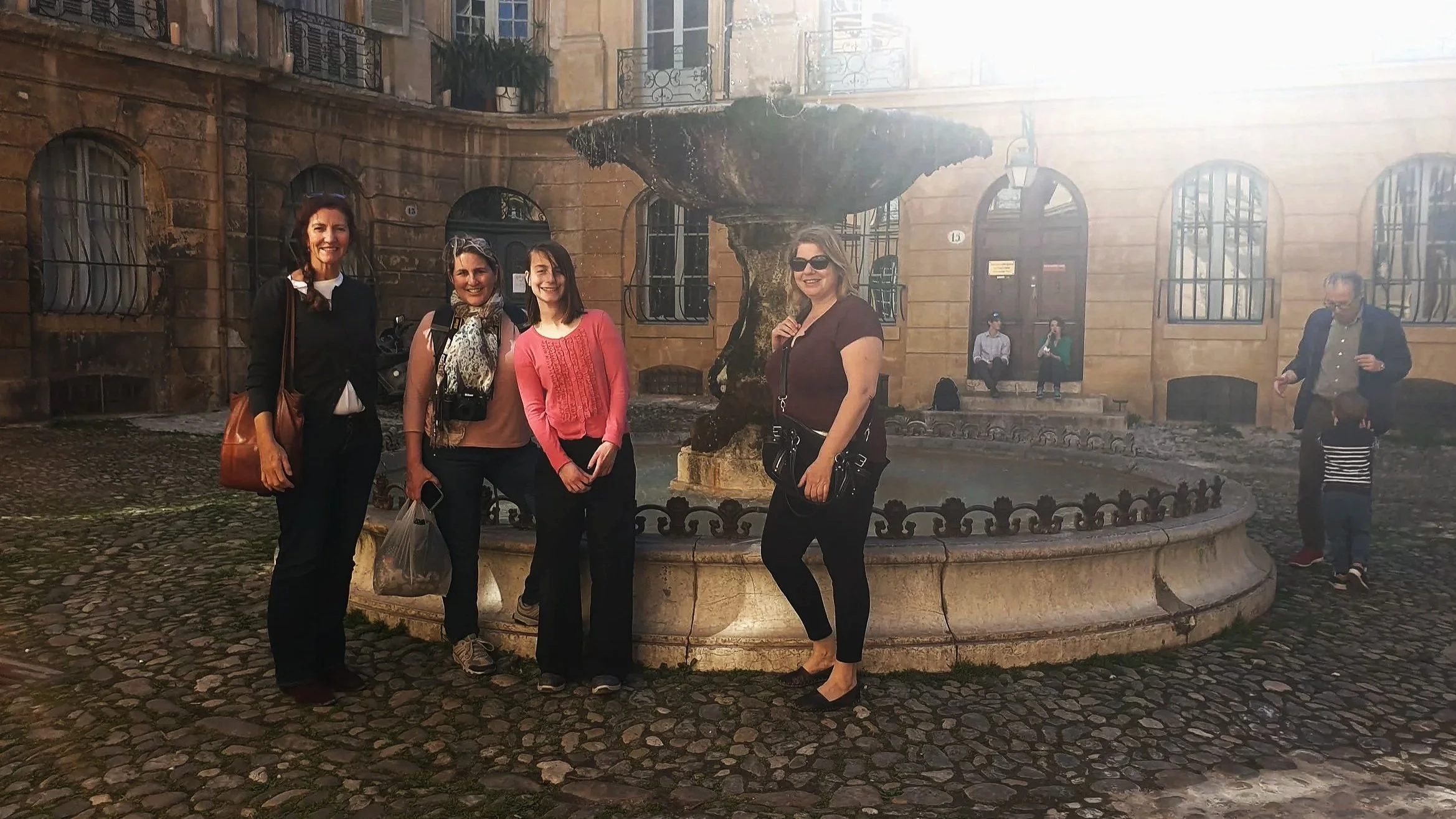 Four women standing in front of a fountain in a cobblestone courtyard, with a historic building in the background, smiling and posing for the photo.