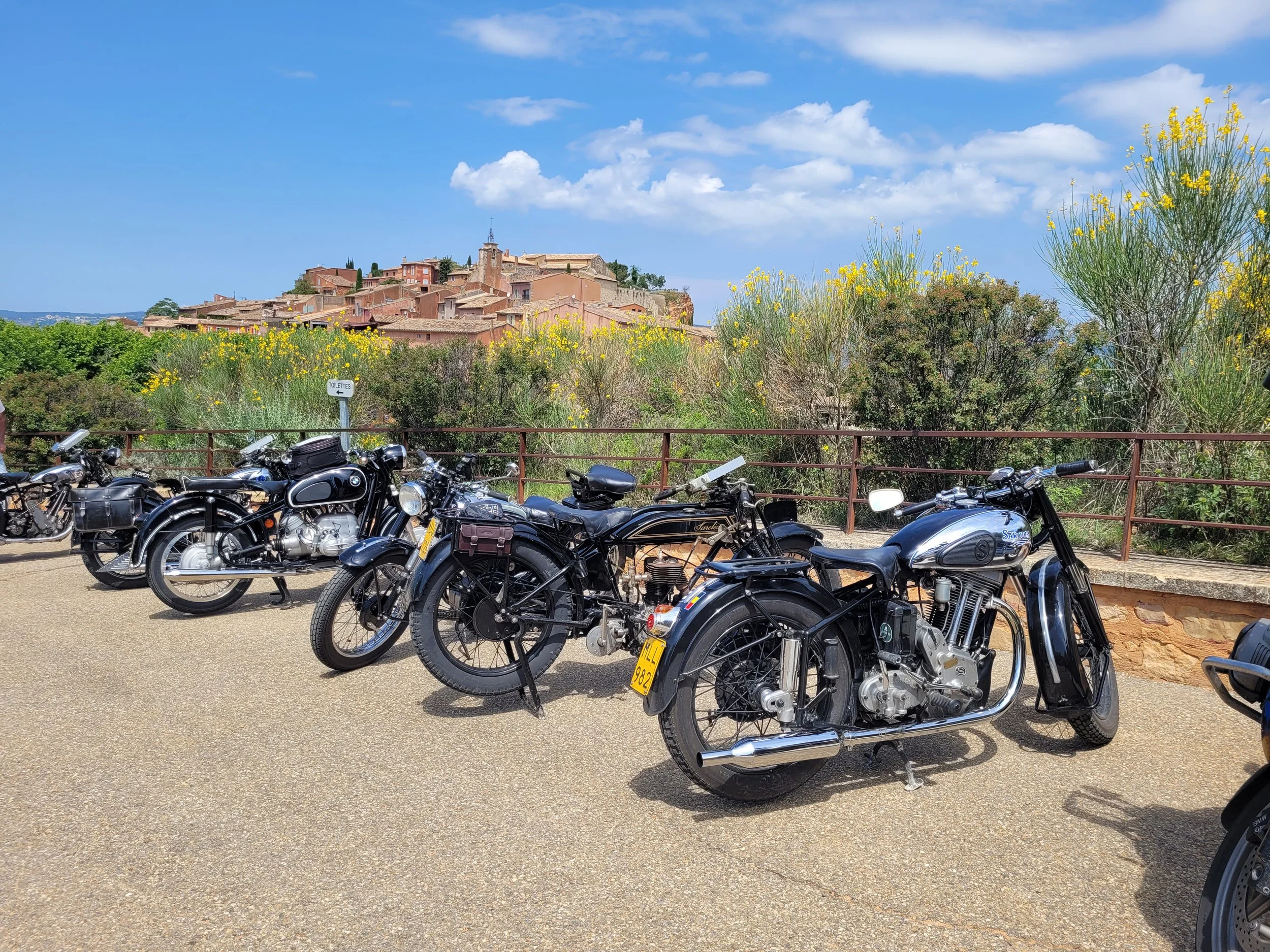 Several vintage motorcycles parked on a paved area, with a scenic hillside town and greenery in the background under a blue sky with clouds.