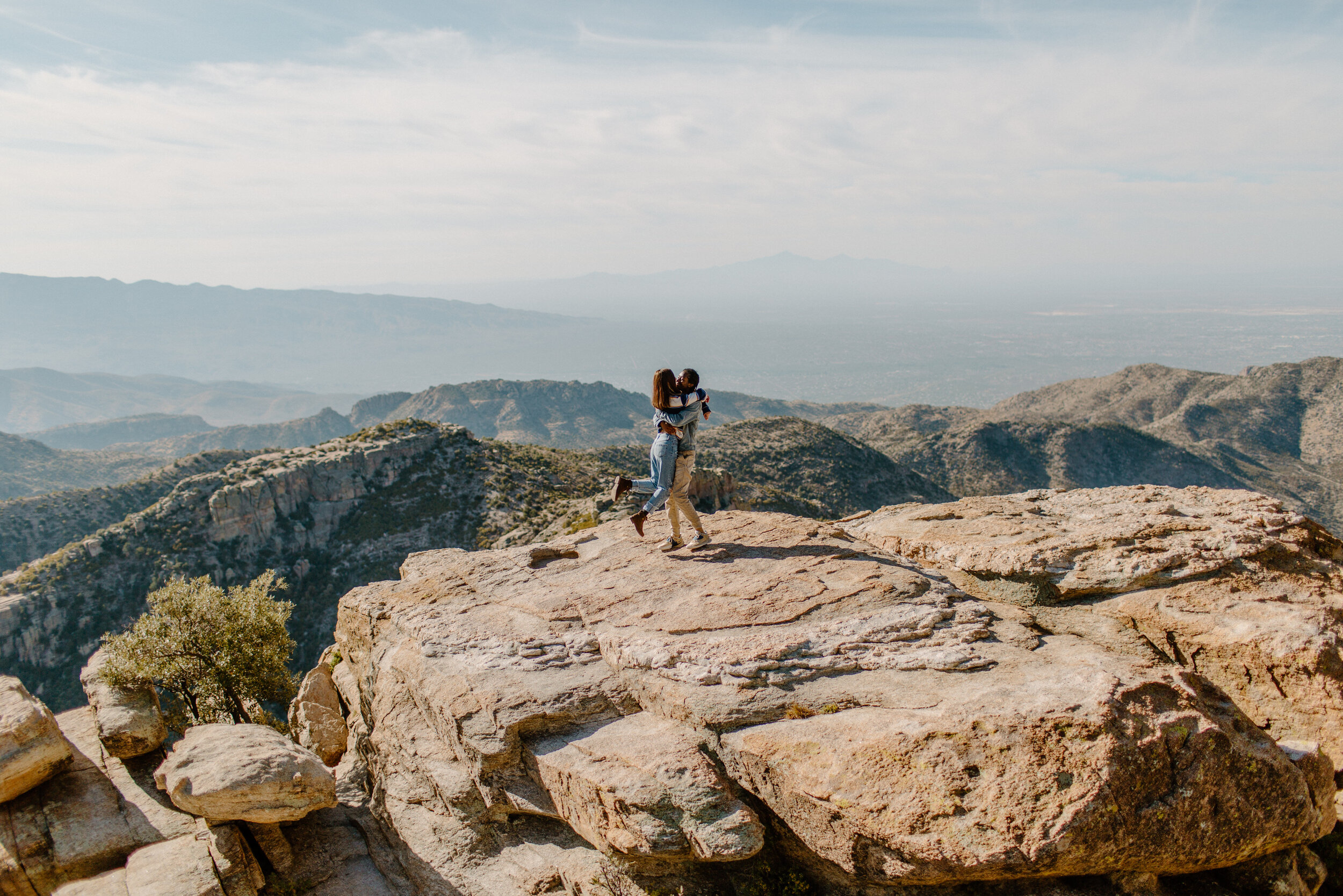  Wide shot of a man holding and lifting his girlfriend up in front of a mountain view at Windy Point on Mount Lemmon in Tucson Arizona. Arizona couples photographer, Lucy B. Photography. 