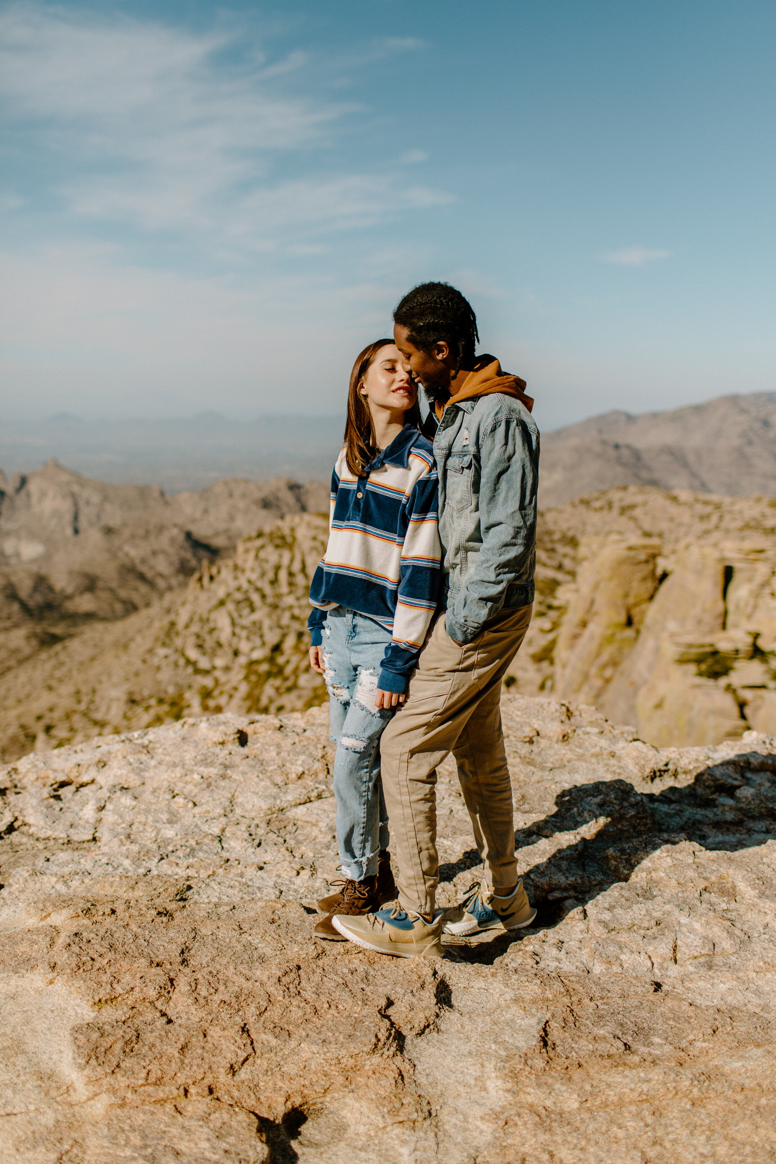  Woman stands in front of her boyfriend and turns her head to kiss him while standing on a rock at Windy Point on Mount Lemmon in Tucson Arizona. Tucson couples photographer, Lucy B. Photography. 