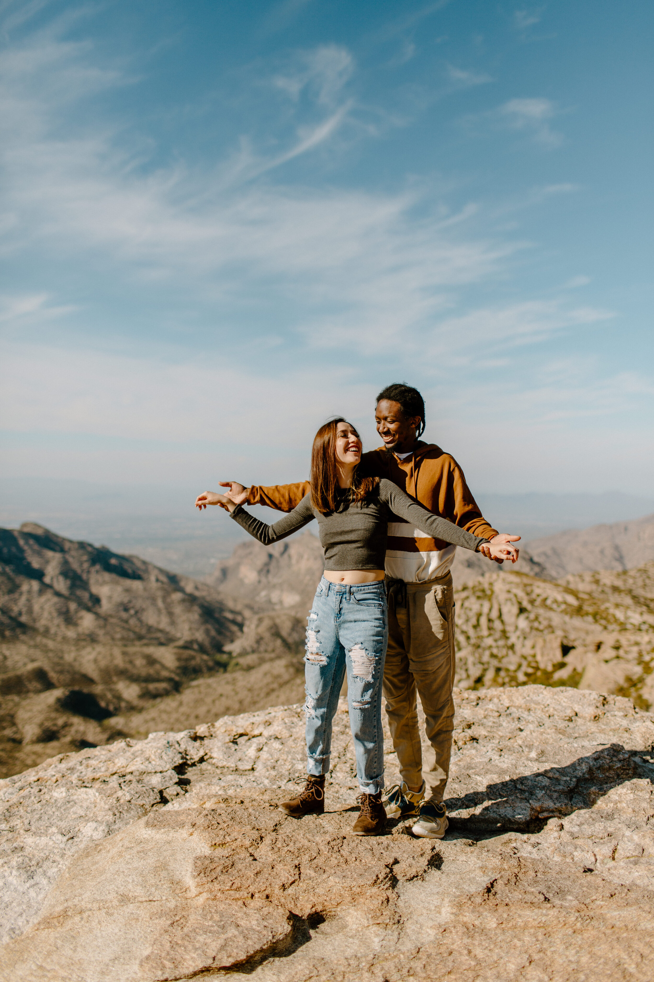  Couple smiles at each other as woman stands in front of her boyfriend and he holds her hands out like an airplane. Mountain view in the background at Windy Point on Mount Lemmon in Tucson Arizona. Arizona couples photographer, Lucy B. Photography. 