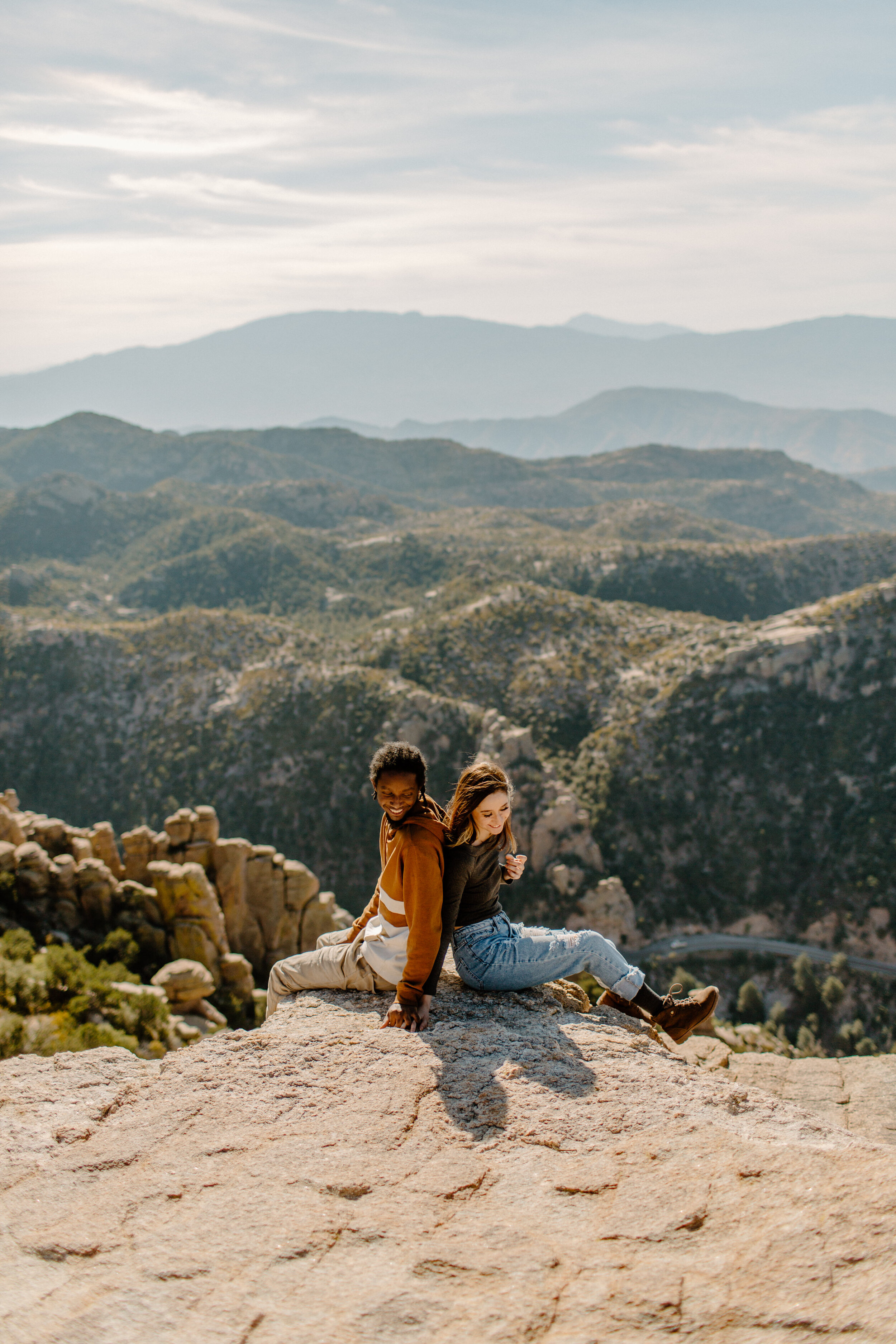  Couple sits back to back and turns to look at each other in front of a mountain view at Windy Point on Mount Lemmon in Tucson Arizona. Tucson couples session, Lucy B. Photography. 