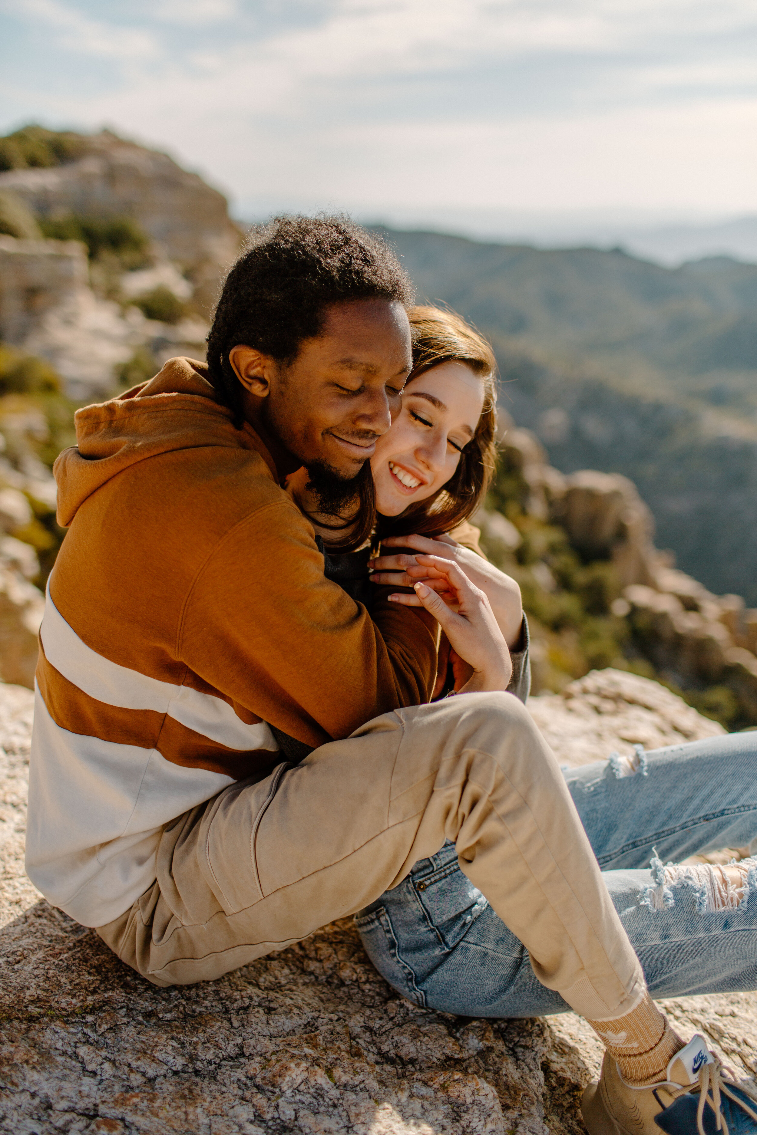  Couple sitting on a rock in front of a mountain view as the man holds his girlfriend close and they both smile at Windy Point on Mount Lemmon in Tucson Arizona. Tucson couples photographer, Lucy B. Photography, 