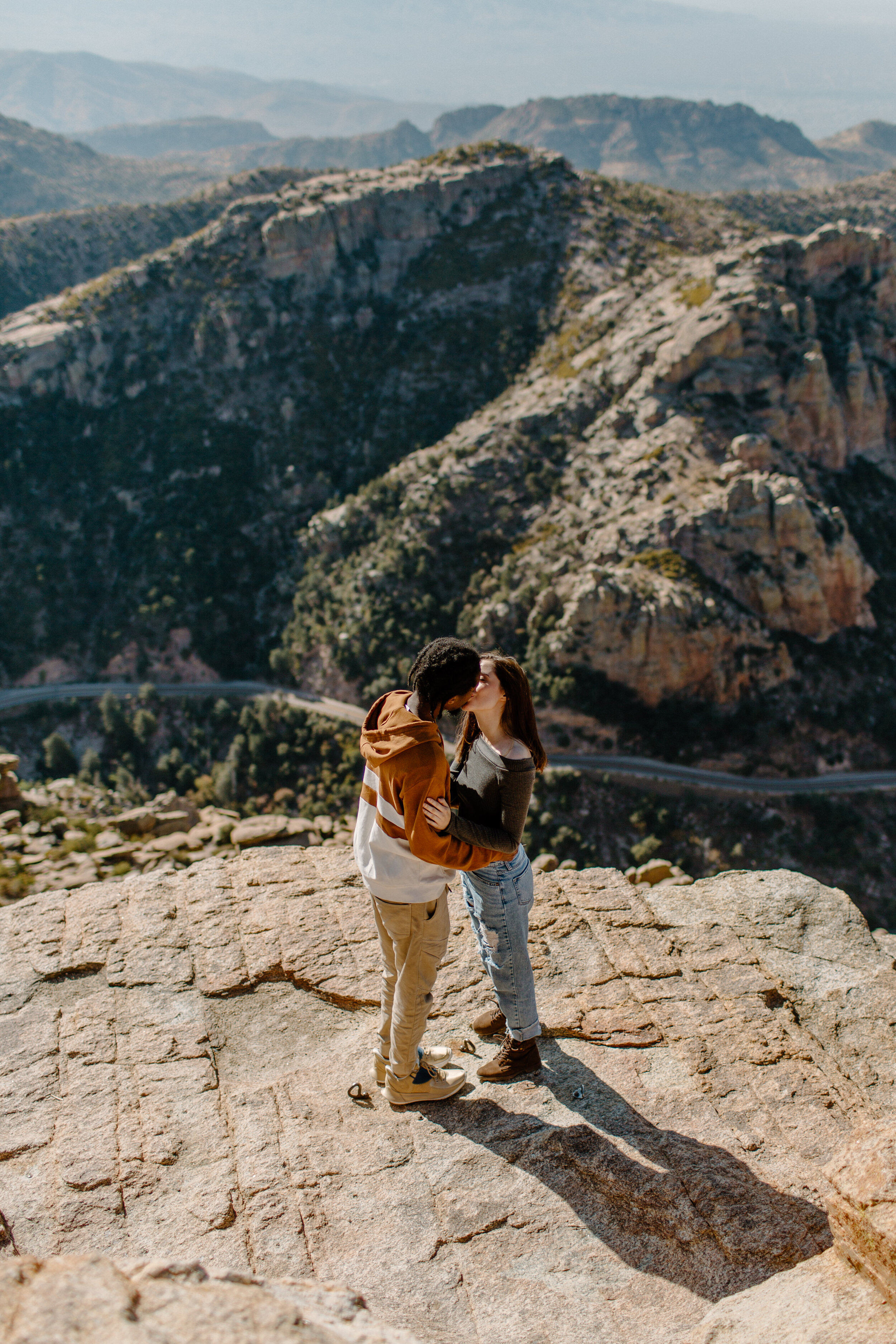  Couple kisses while standing on a rock with a view of a mountain road in the background at Windy Point on Mount Lemmon in Tucson Arizona. Arizona couples photography, Lucy B. Photography. 