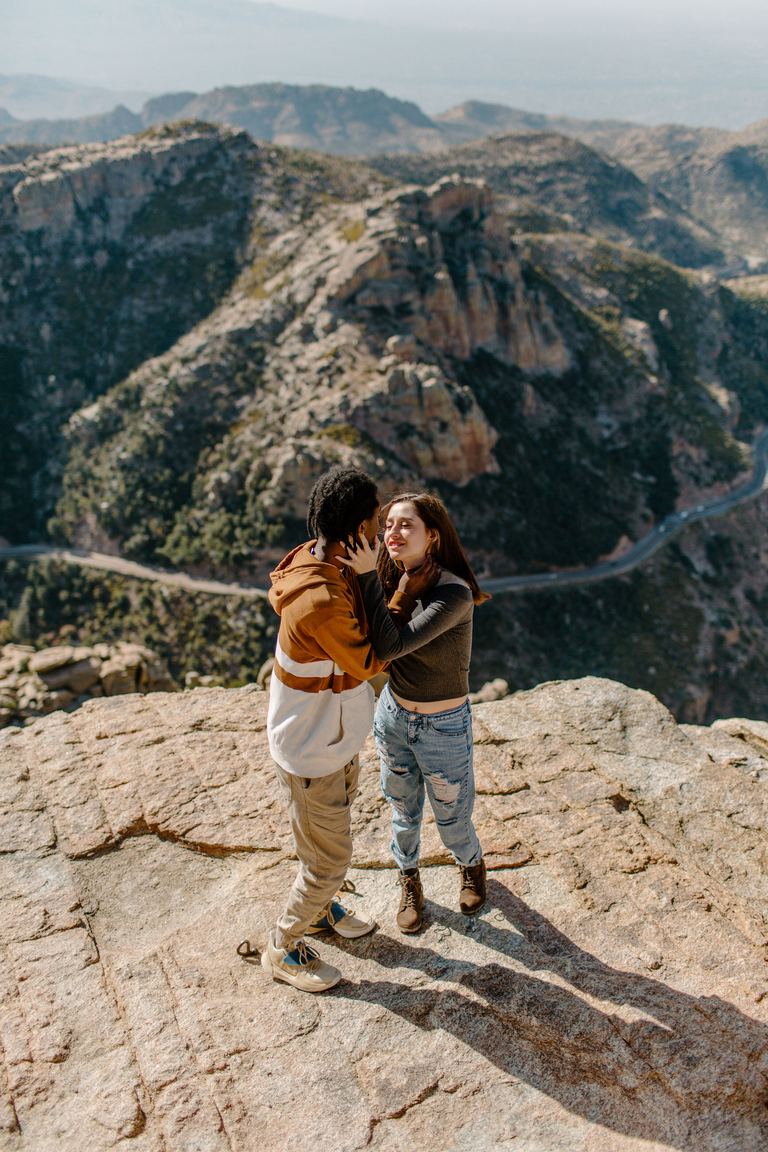  Woman holds her boyfriend’s face and smiles at him while standing on a rock with a view of a mountain road behind them at Windy Point on Mount Lemmon in Tucson Arizona. Tucson couples photography, Lucy B. Photography. 