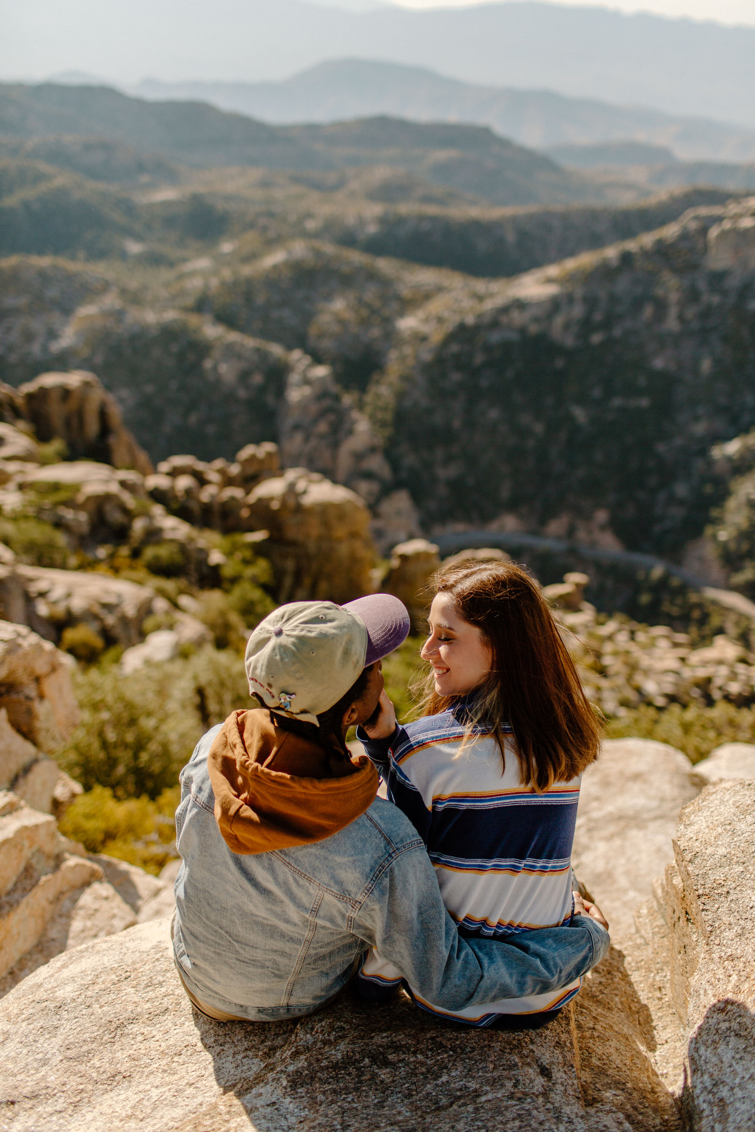  Couple sits on a rock and smiles at each other in front of a beautiful mountain view at Windy Point on Mount Lemmon in Tucson Arizona. Tucson couples photographer, Lucy B. Photography. 