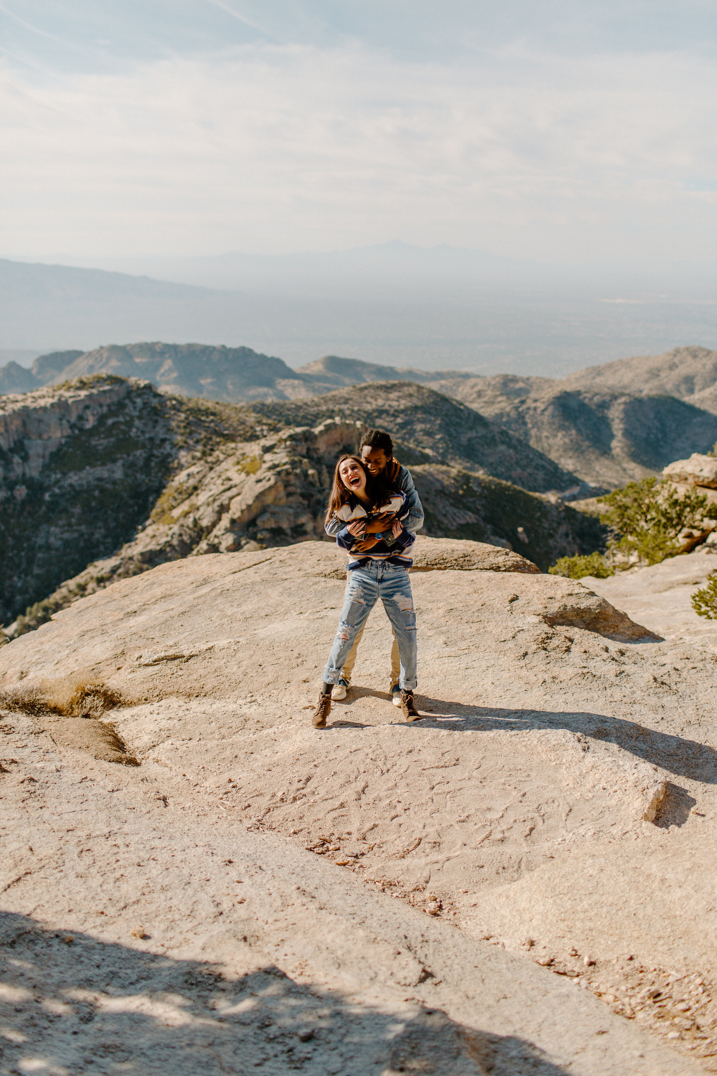  Boyfriend stands behind his girlfriend and holds her while they both laugh at Windy Point on Mount Lemmon in Tucson Arizona. View of mountains in the back. Arizona couples photographer, Lucy B. Photography. 