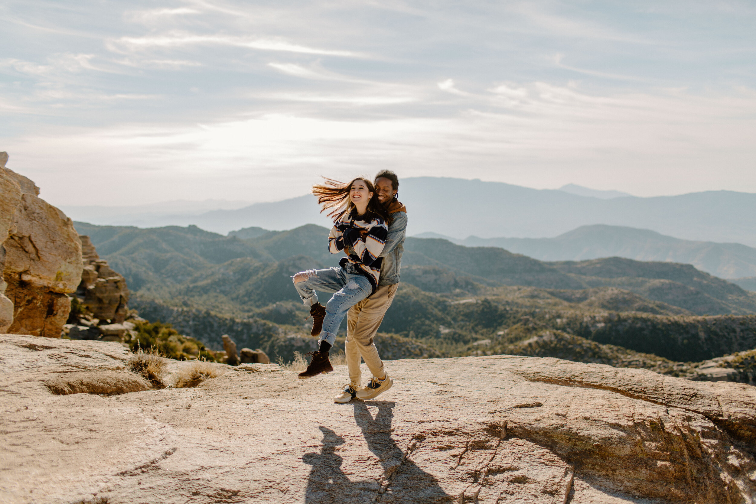  Man holding his girlfriend and twirling her around as she laughs and her hair flies in the wind. View of mountains in the background at Windy Point on Mount Lemmon in Tucson Arizona. Tucson couples photographer, Lucy B. Photography. 