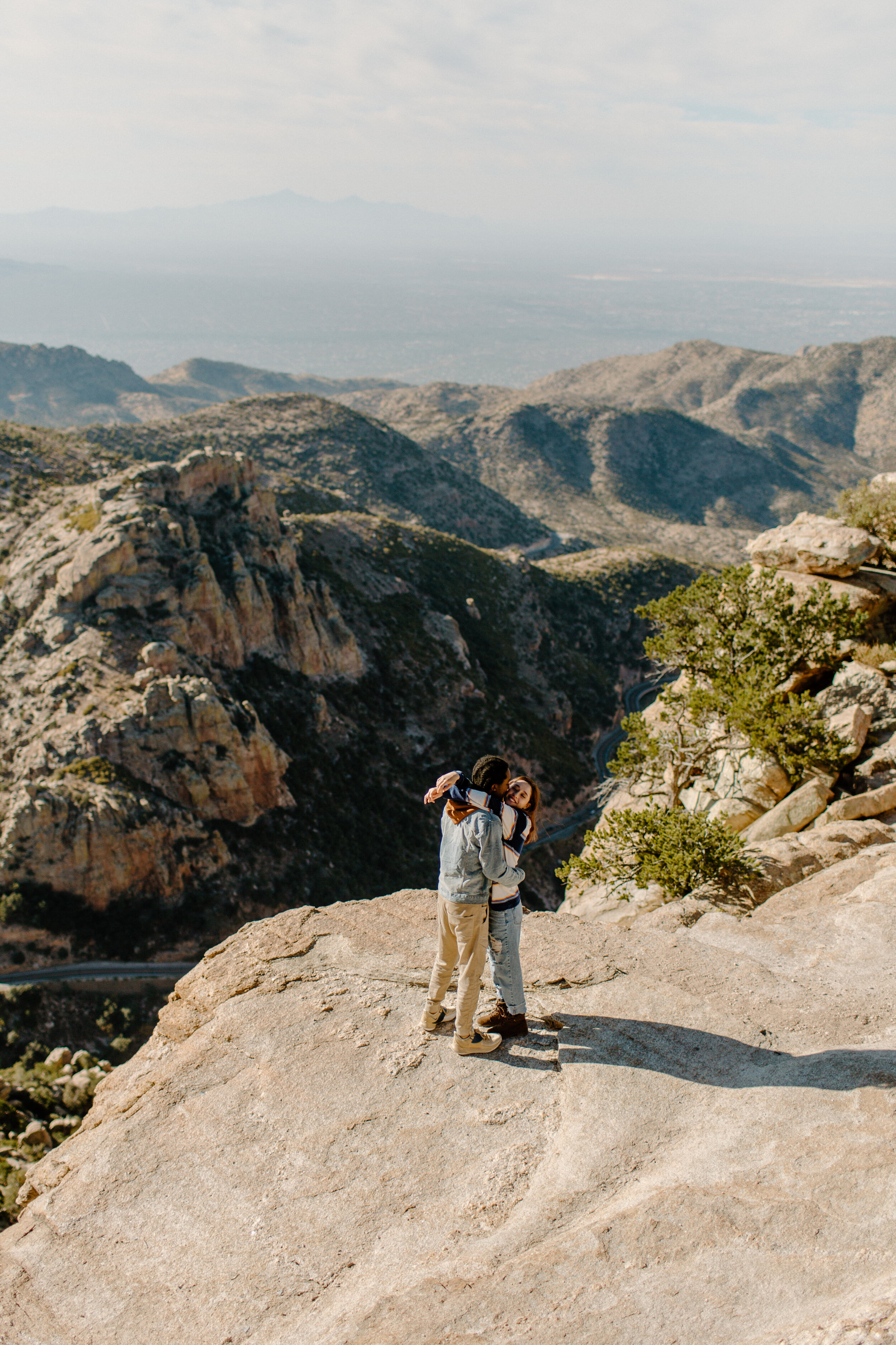  Photo looks down at a couple standing and snuggling in front of a mountain view at Windy Point on Mount Lemmon in Tucson Arizona. Tucson couples photographer, Lucy B. Photography. 