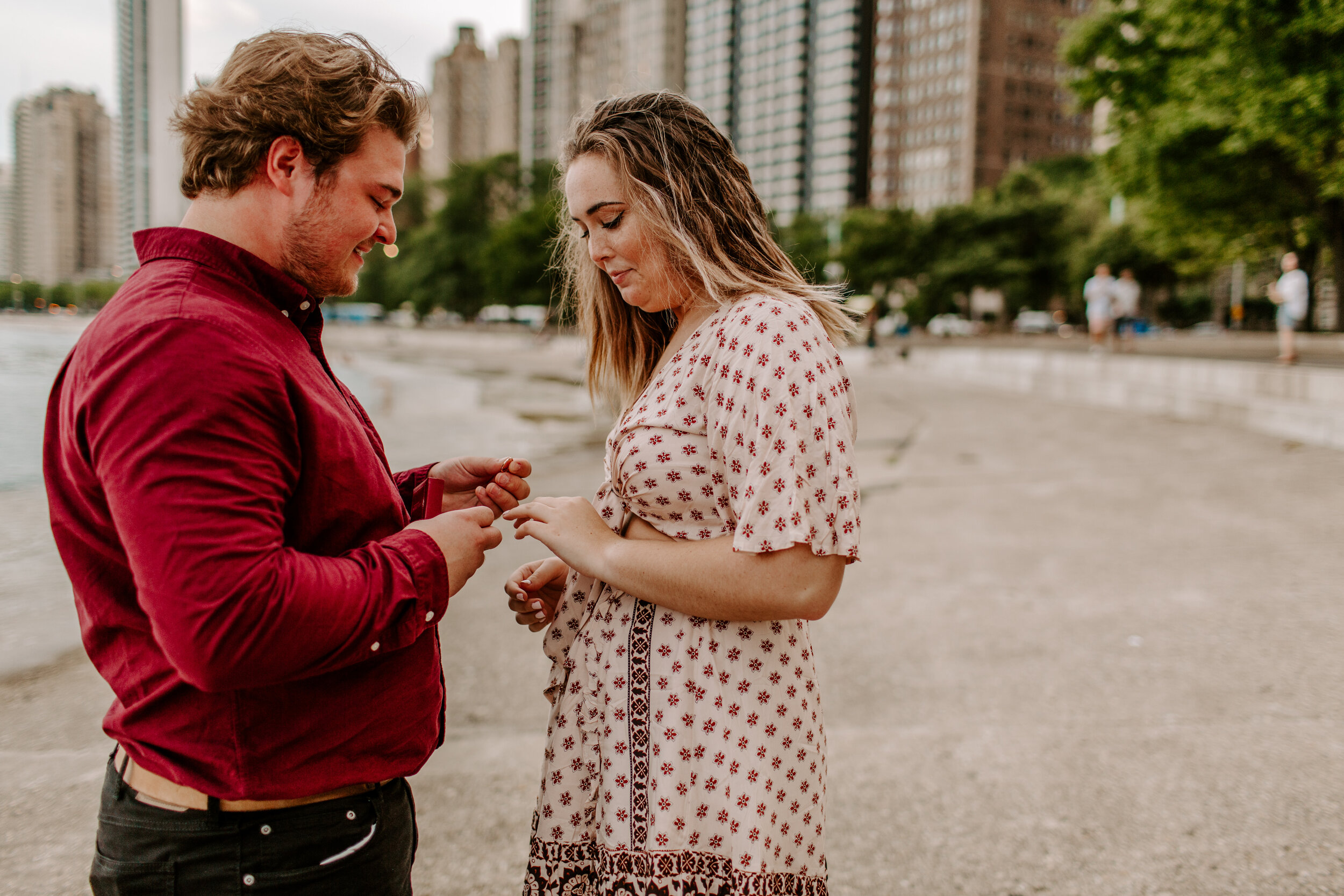  Man puts ring on his new fiance’s finger after a surprise proposal at North Avenue Beach in Chicago next to Lake Michigan with the Chicago skyline in the back. Lucy B. Photography. 
