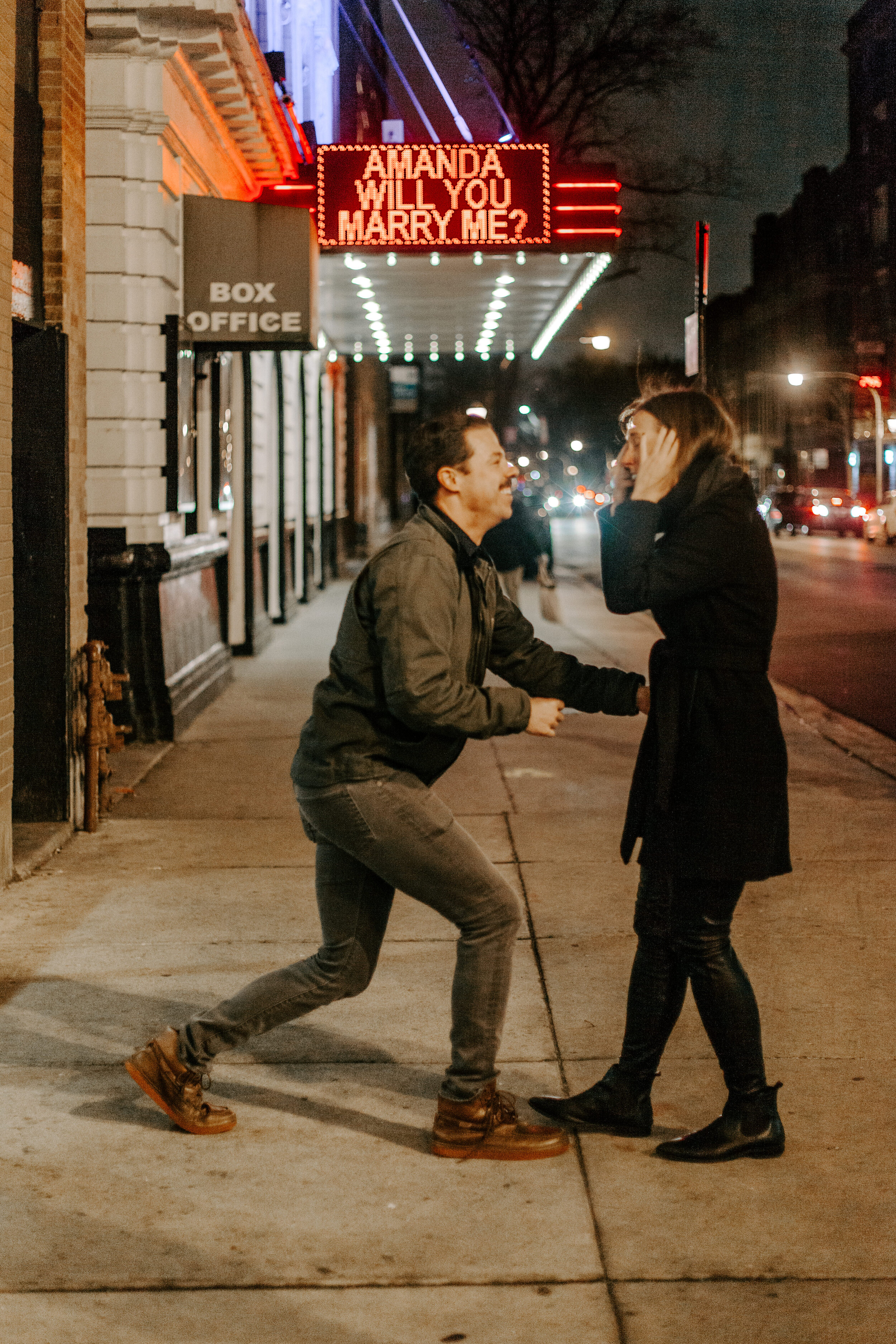  Chicago surprise proposal, man stands up from one knee after proposing as his fiance reacts emotionally in front of theater marquee reading Amanda Will You Marry Me? Chicago engagement photographer, Lucy B. Photography 