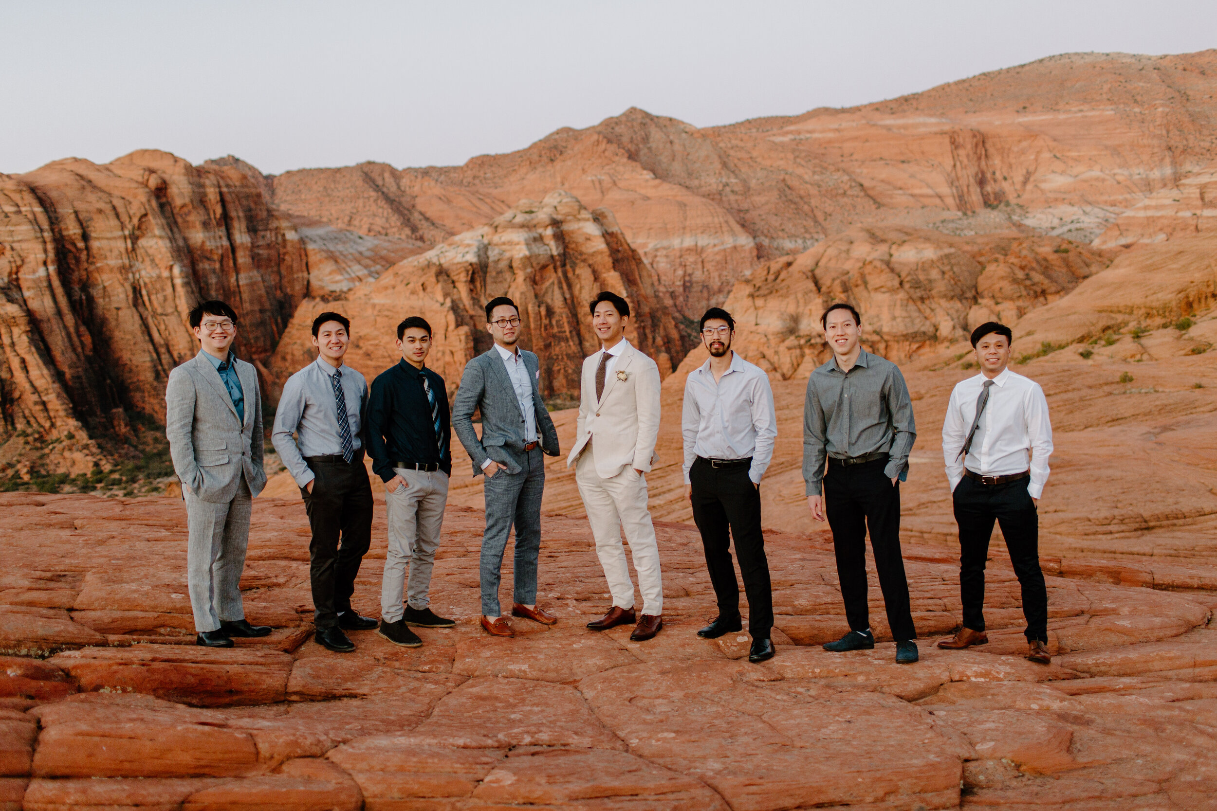  Groom and his groomsmen stand in a line in front of red rocks and smile at the camera at Snow Canyon State Park in St. George Utah. Utah elopement photographer, Lucy B. Photography. 