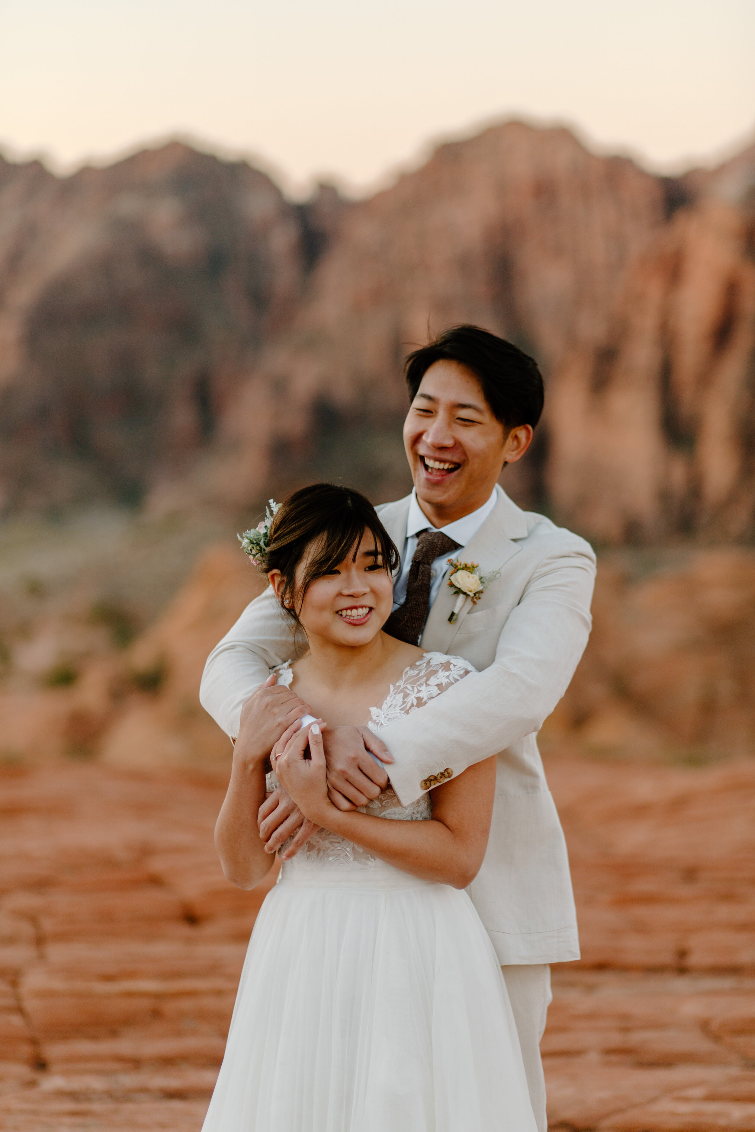  Groom stands behind his bride and holds her as they laugh at Snow Canyon State Park in St. George Utah. Utah elopement photographer, Lucy B. Photography. 