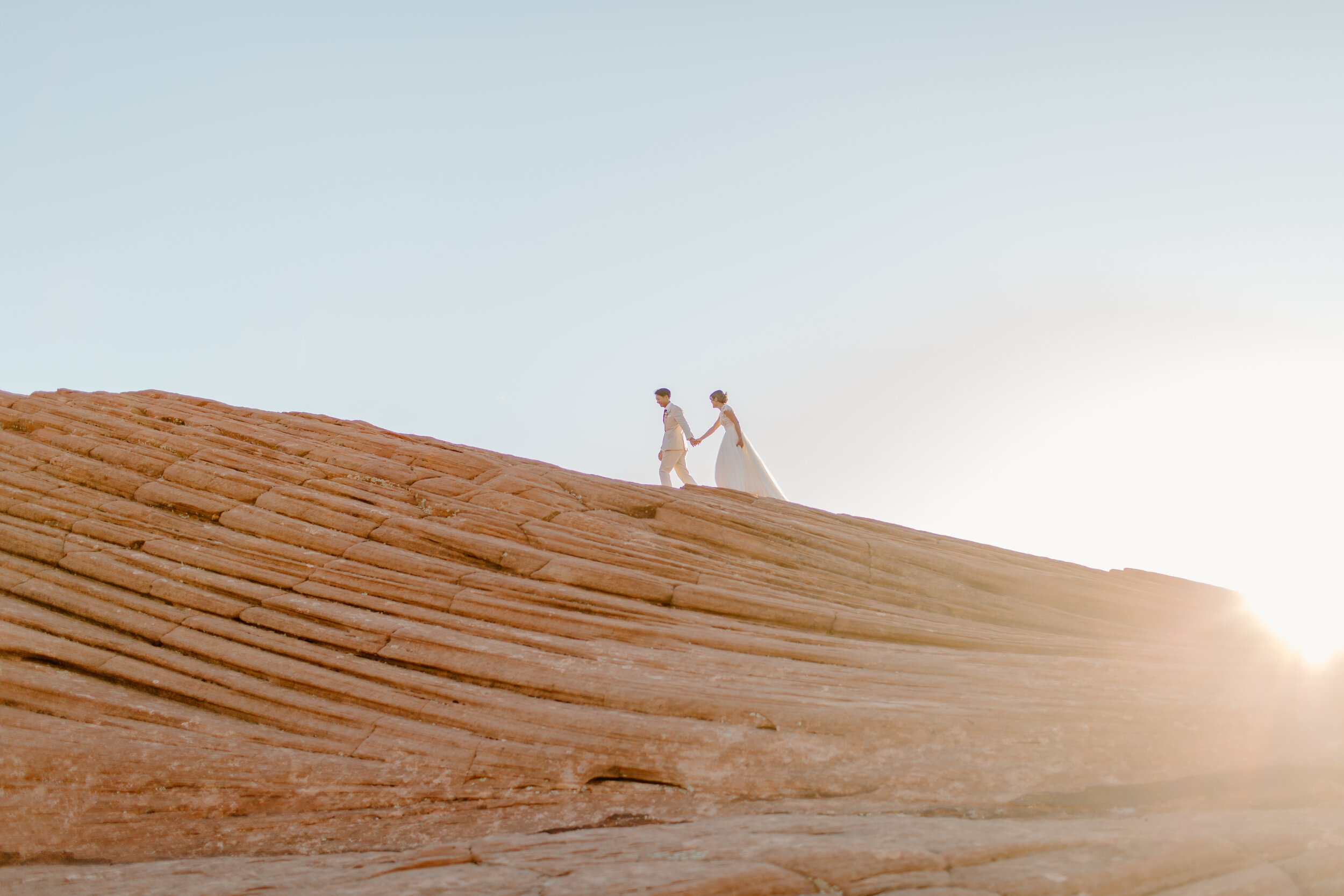  Wide shot of a Utah elopement couple holding hands and walking along a ridge with the sun behind them at Snow Canyon State Park in St. George Utah. Utah elopement photographer, Lucy B. Photography. 
