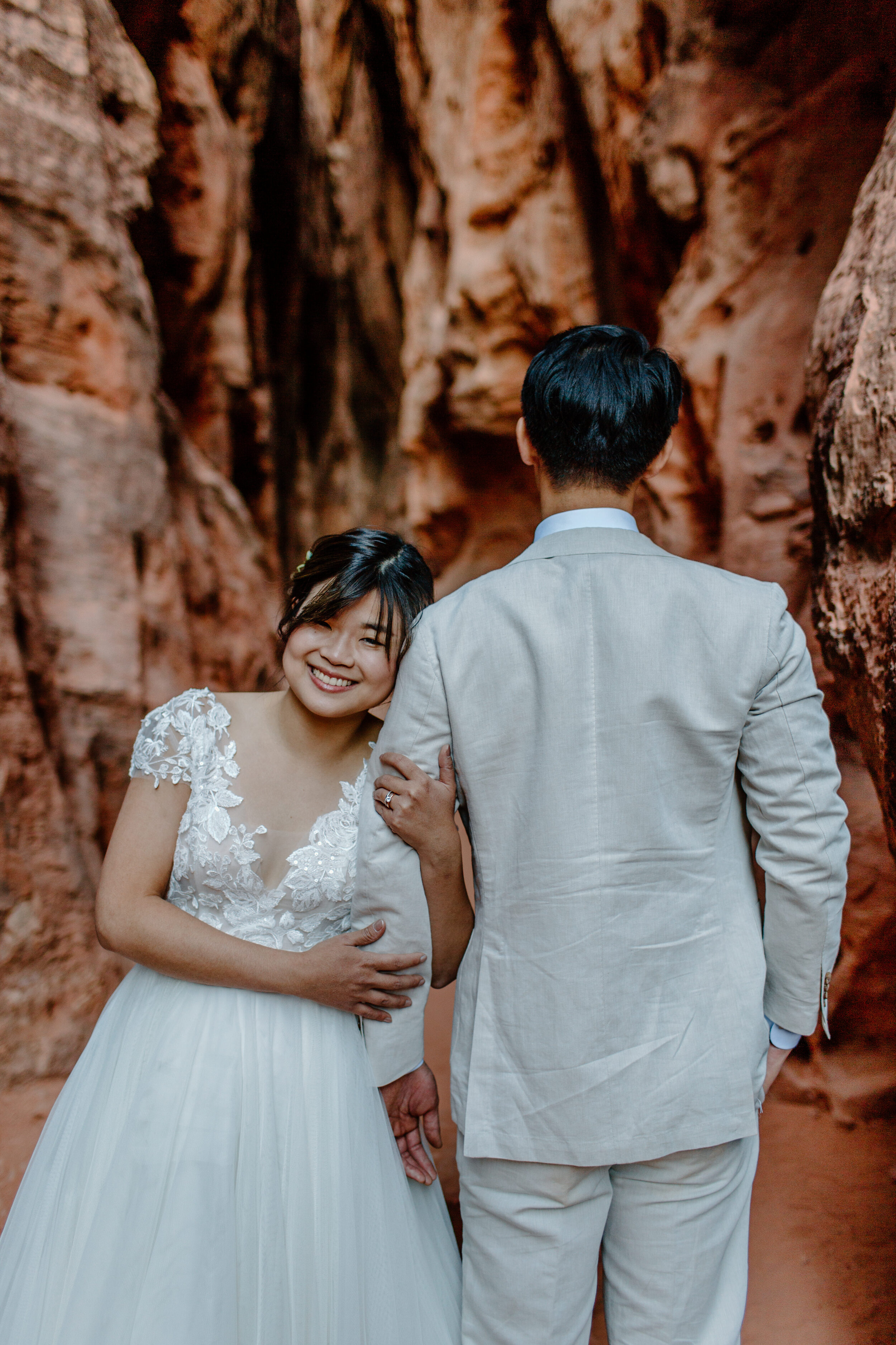  Utah elopement couple stands in a slot canyon at Snow Canyon State Park in St. George Utah. Groom faces away while bride holds his arm and looks at the camera. Utah elopement photographer, Lucy B. Photography. 