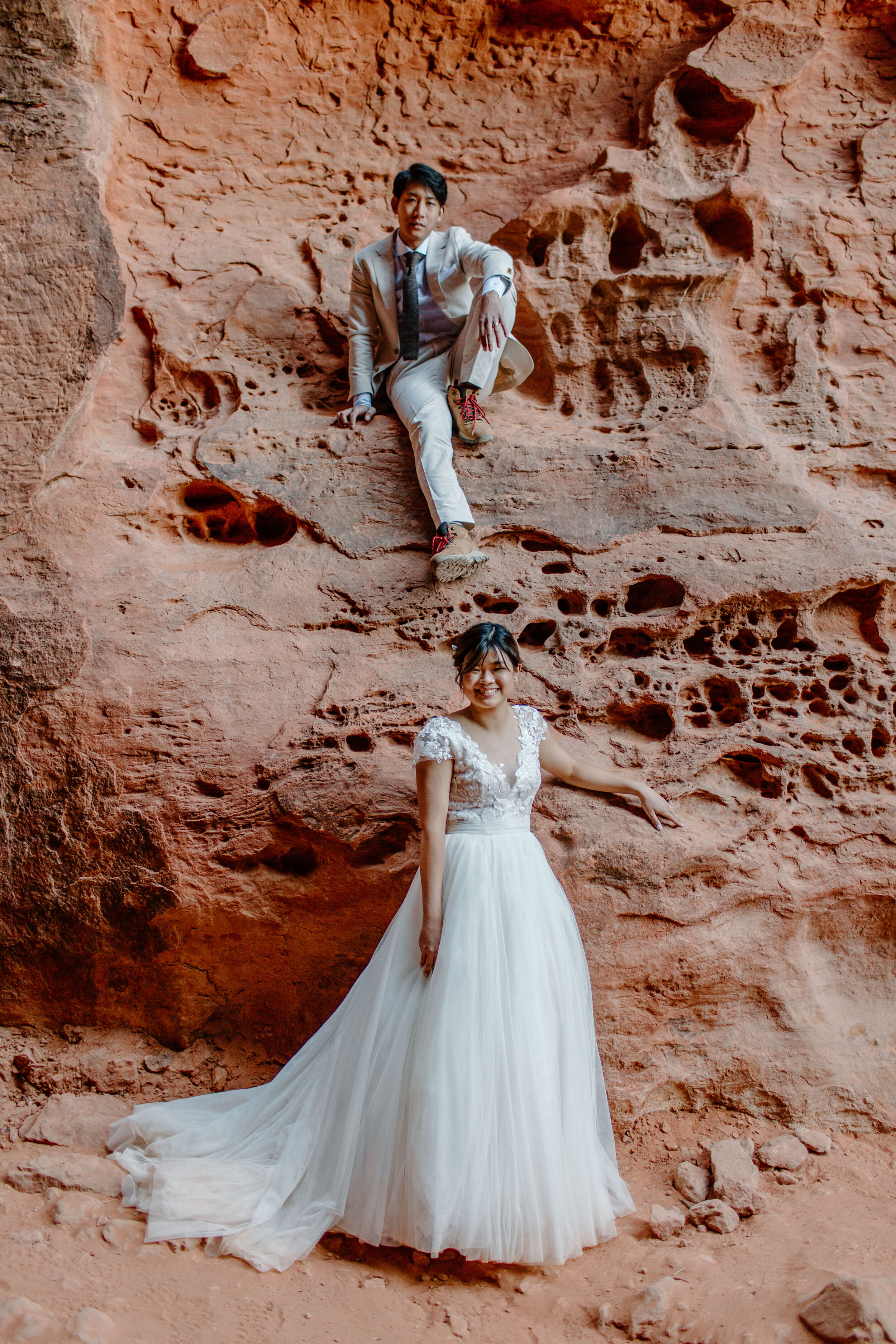  Utah elopement groom sits on a ledge on the side of a slot canyon while the bride stands beneath him and they smile at the camera at Snow Canyon State Park in St. George Utah. Utah elopement photographer, Lucy B. Photography. 