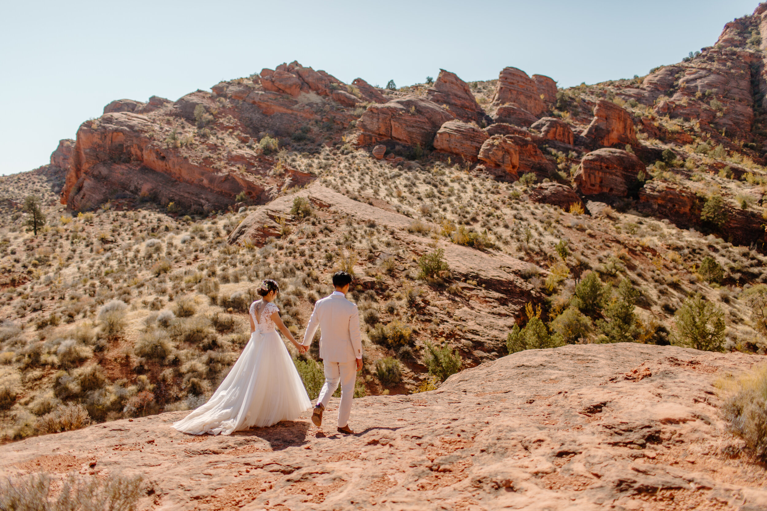  Wide shot of a Utah elopement couple hold hands and walk towards a mountain landscape in St. George Utah. Utah elopement photographer, Lucy B. Photography. 
