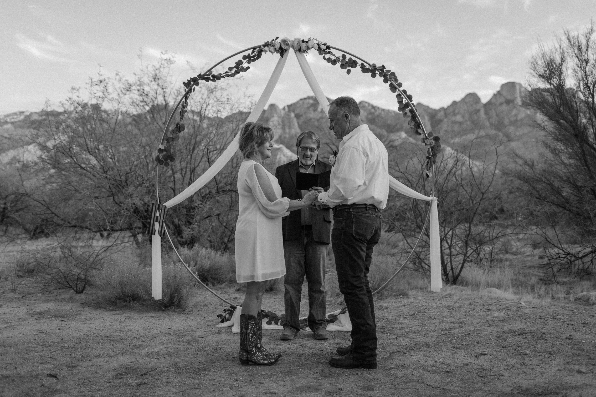  Black and white photo of bride putting a wedding band on her groom’s finger with a mountain backdrop in Catalina State Park in Tucson. Tucson elopement photographer, Lucy B. Photography. 