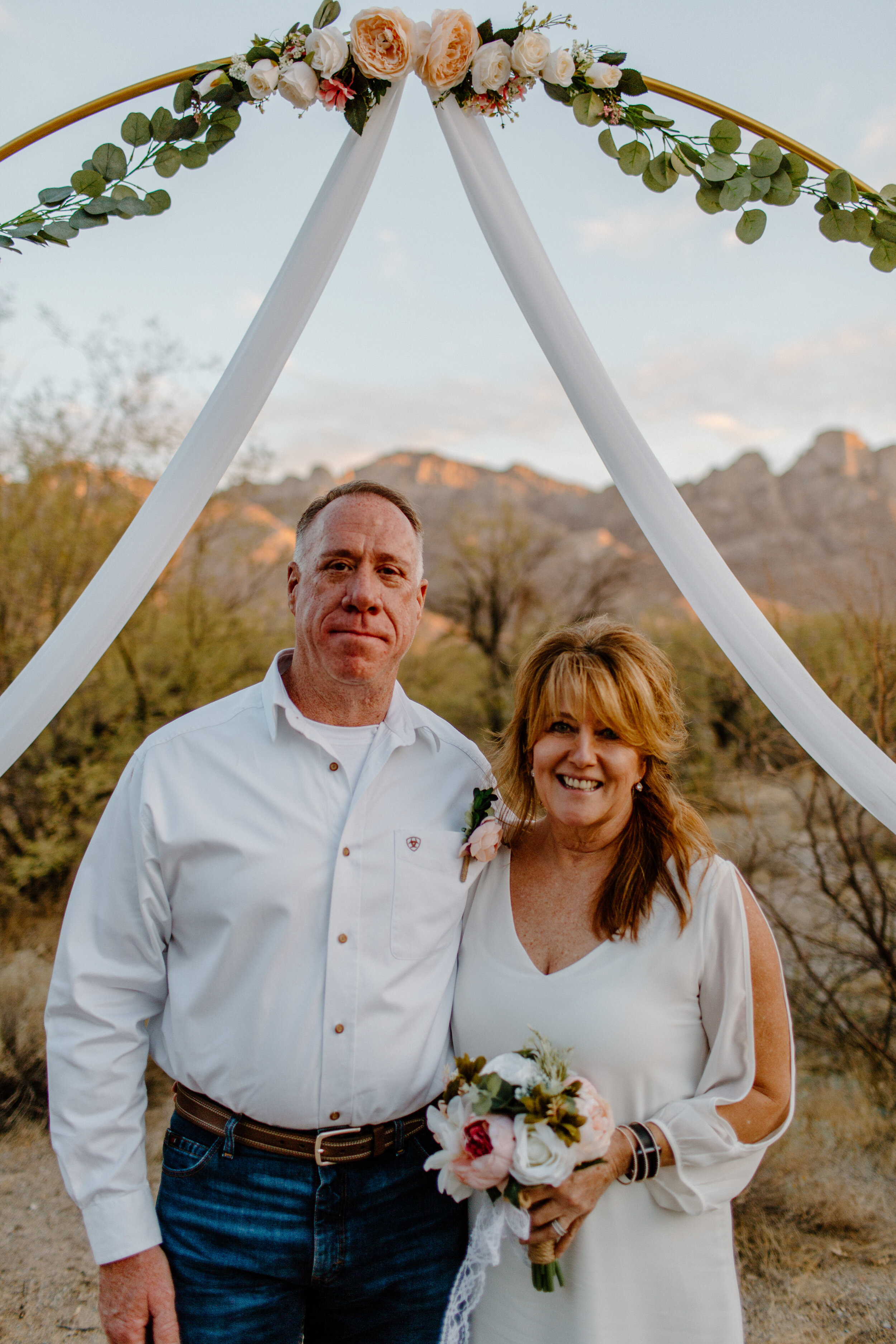  Bride and groom smile for the camera under their floral arch with a mountain backdrop in Catalina State Park in Tucson. Tucson elopement photographer, Lucy B. Photography. 