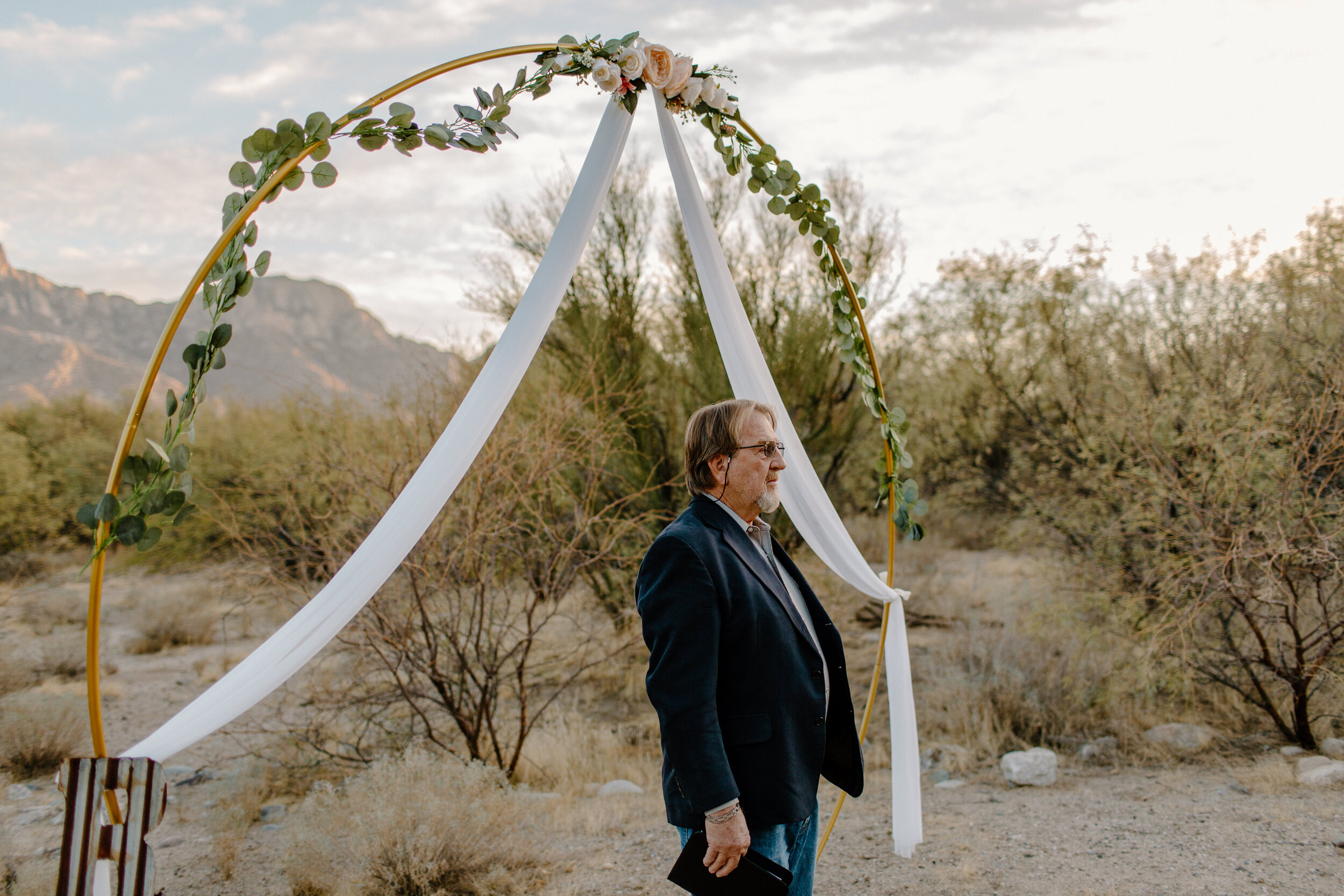  Officiant stands under floral arch and watches the elopement couple walk up in Catalina State Park in Tucson. Tucson elopement photographer, Lucy B. Photography. 