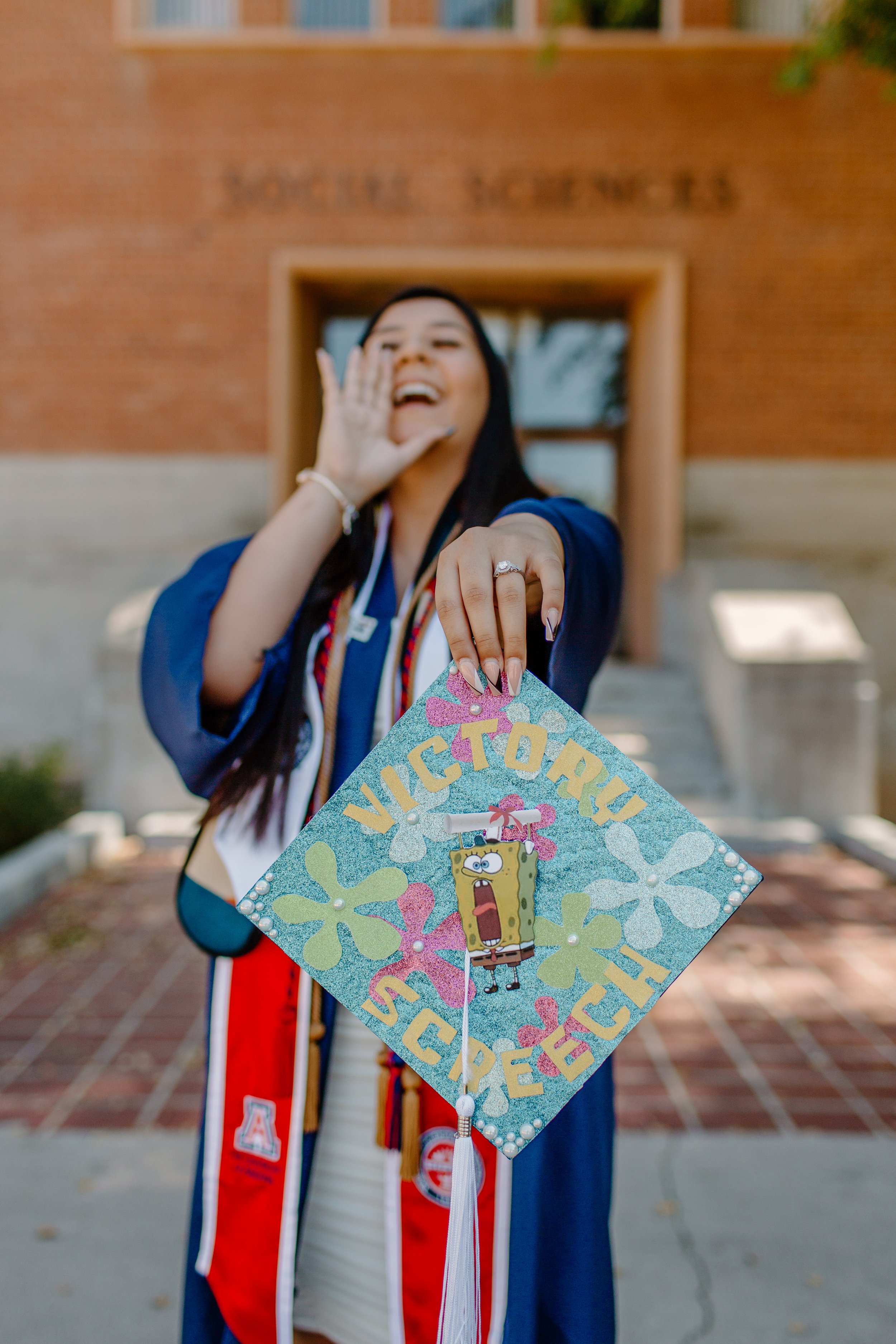 College graduate holds out her decorated cap and mimics a happy scream in the background 