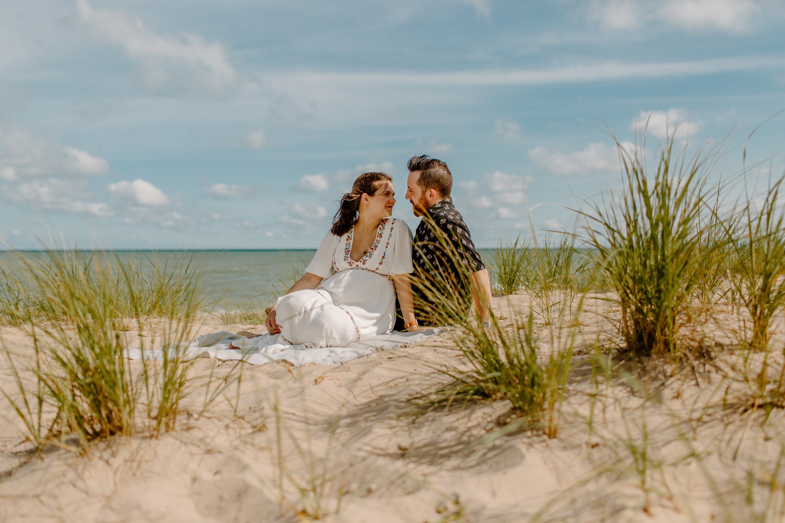  Pregnant couple sits on a towel in the sand in front of Lake Michigan at the Indiana Dunes 