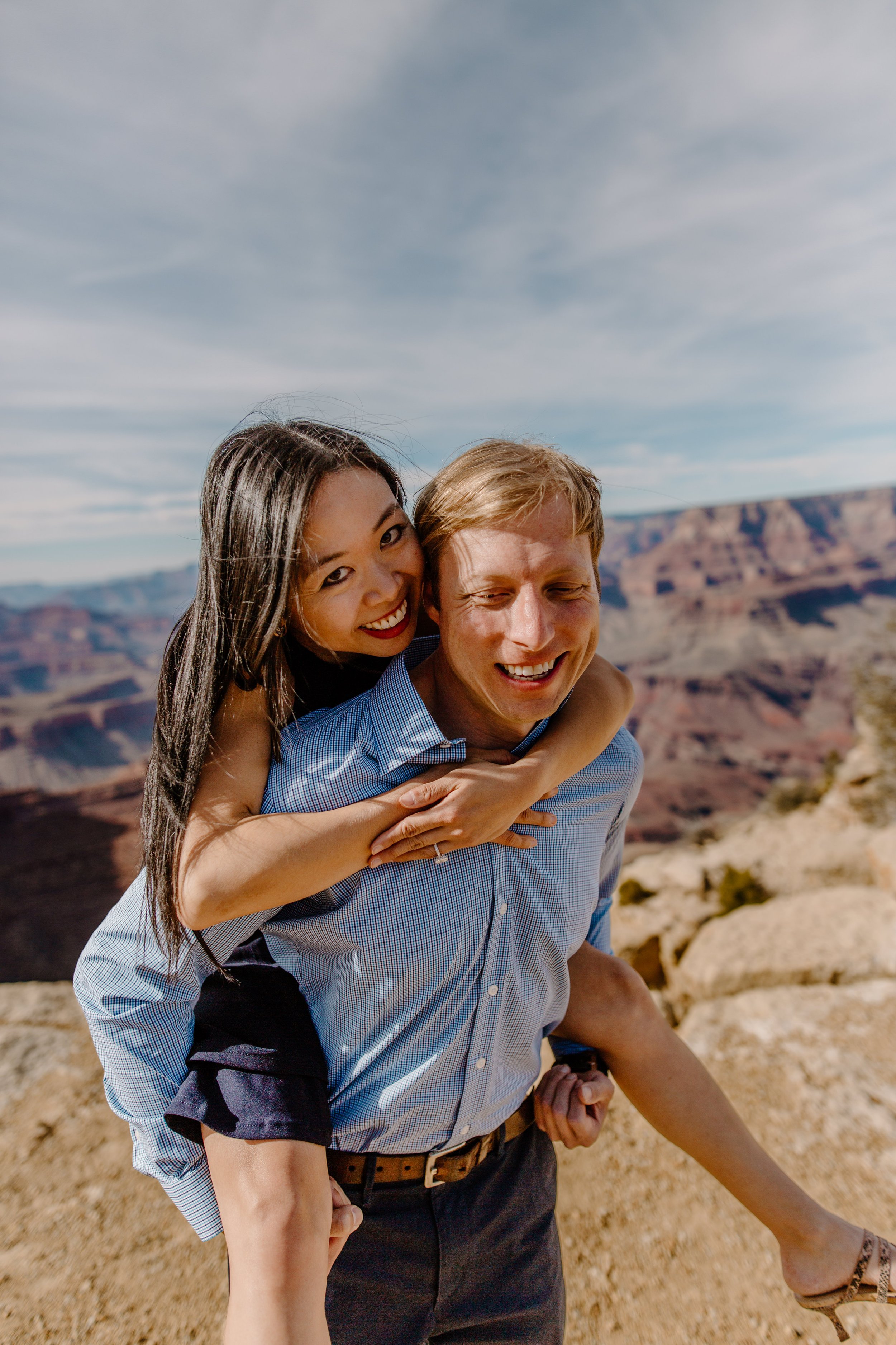  A woman on her fiance’s back and they both smile at the camera with the Grand Canyon in the background 