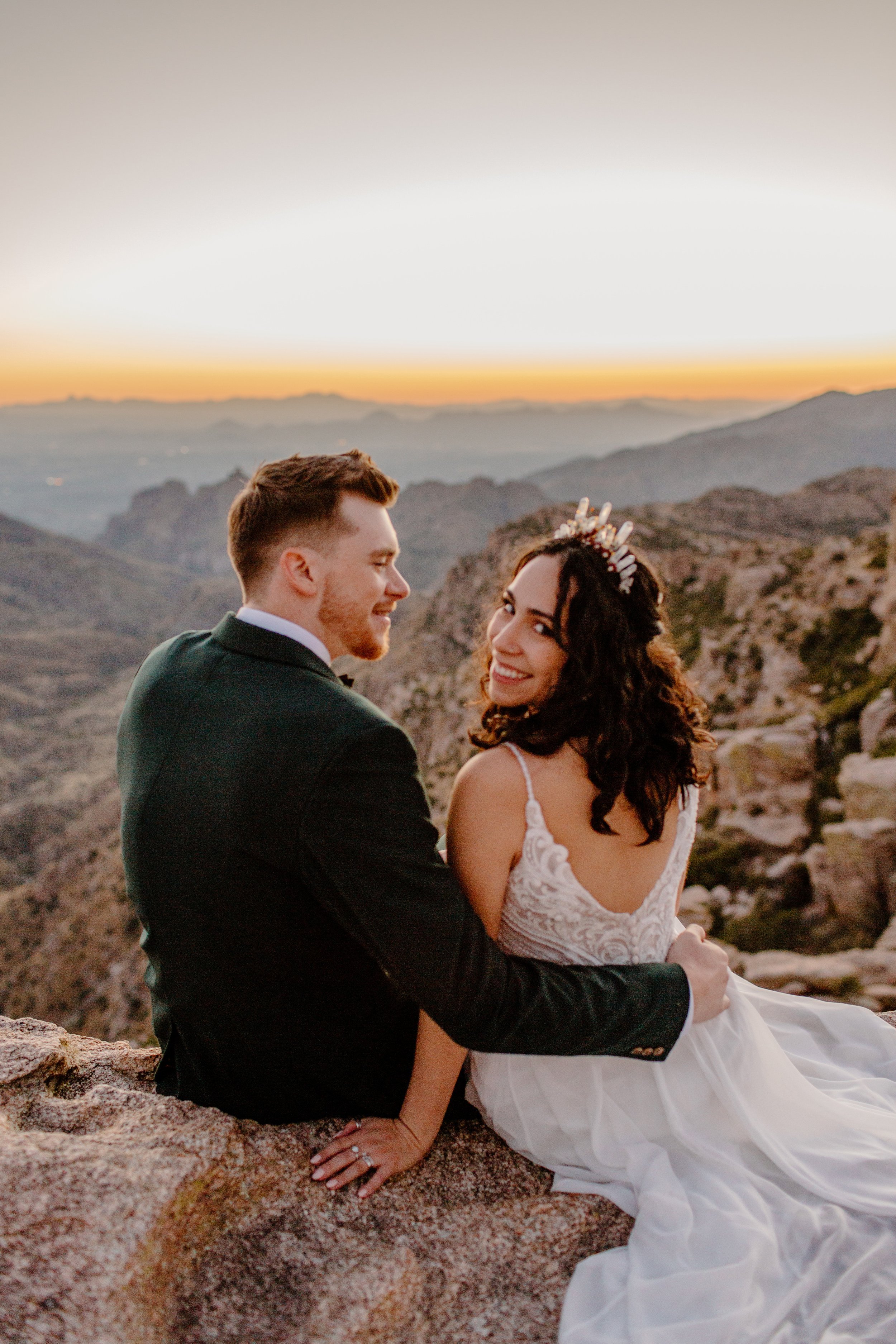  Couple sits on a cliff edge on Mount Lemmon in Tucson Arizona, bride looks back at camera while groom looks at bride 