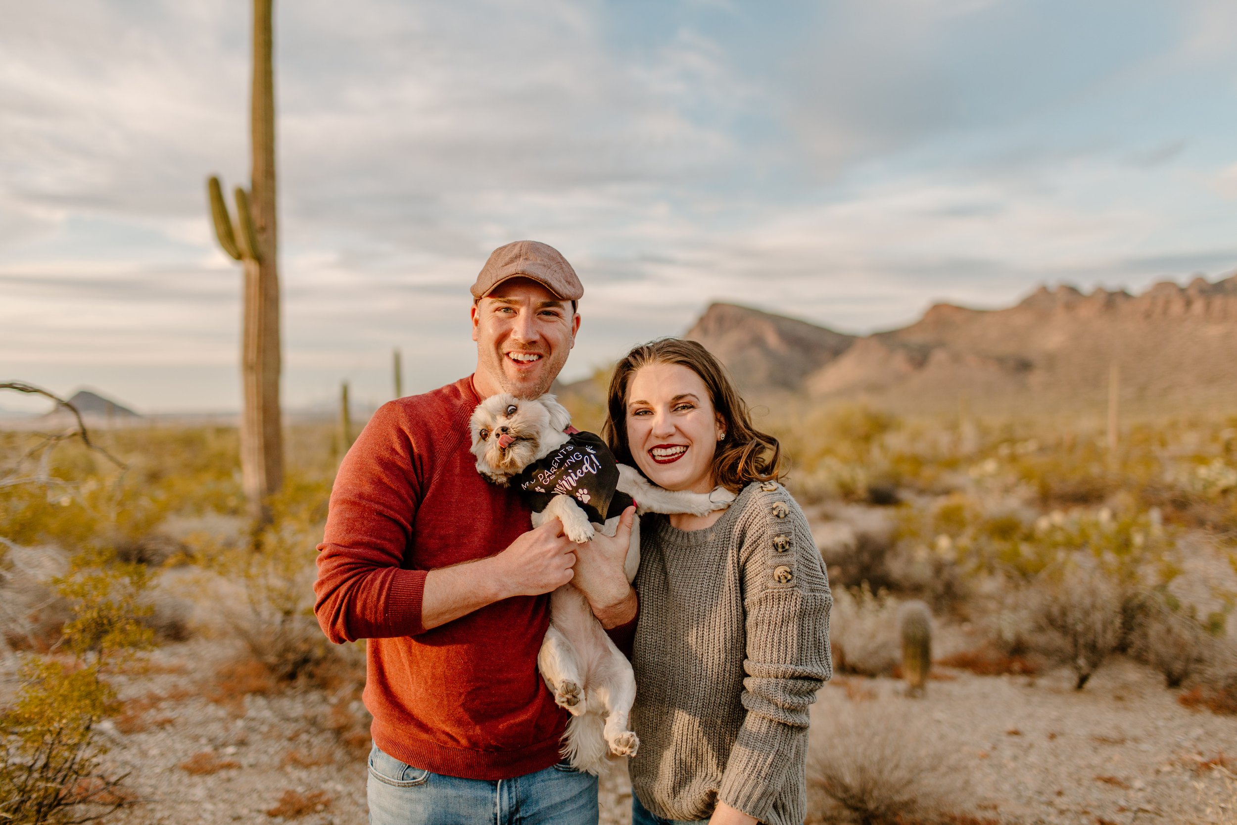  Couple smiles at camera while holding small dog making a silly face in Saguaro National Park in Tucosn Arizona 