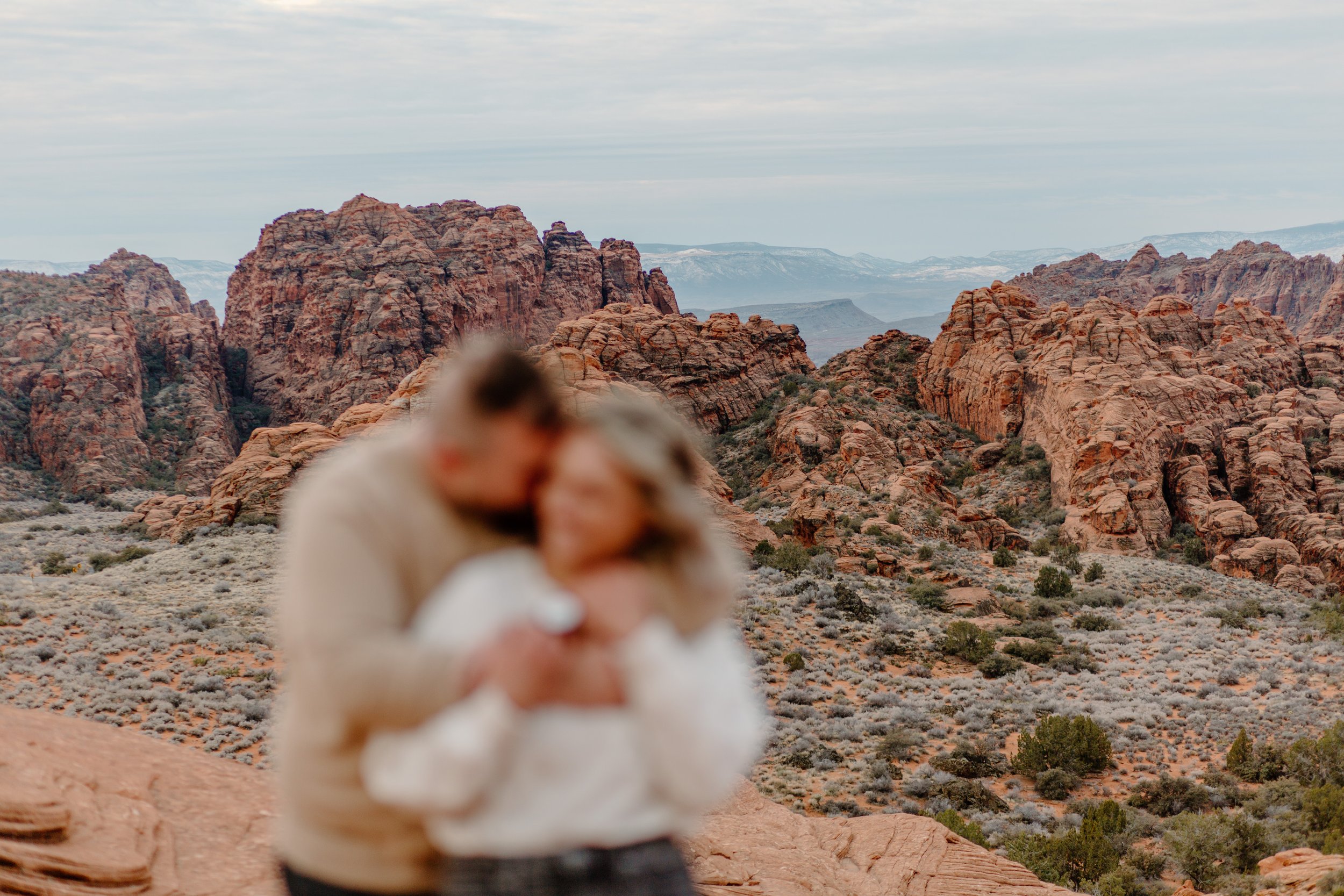  Blurry couple cuddling in foreground while mountains in background are in focus at Snow Canyon State Park in St George Utah 