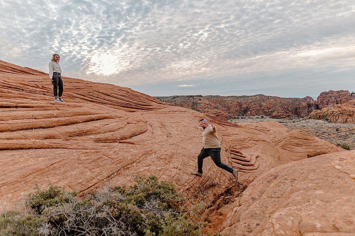  man jumping over the red rocks in Utah with fiancé standing up on cliff 