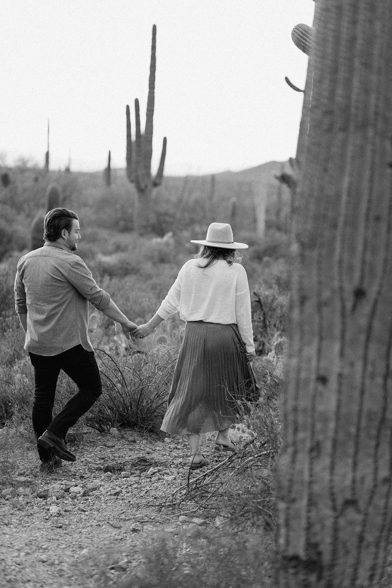  couple walks together holding hands by cactus for Arizona Couples Photographer 