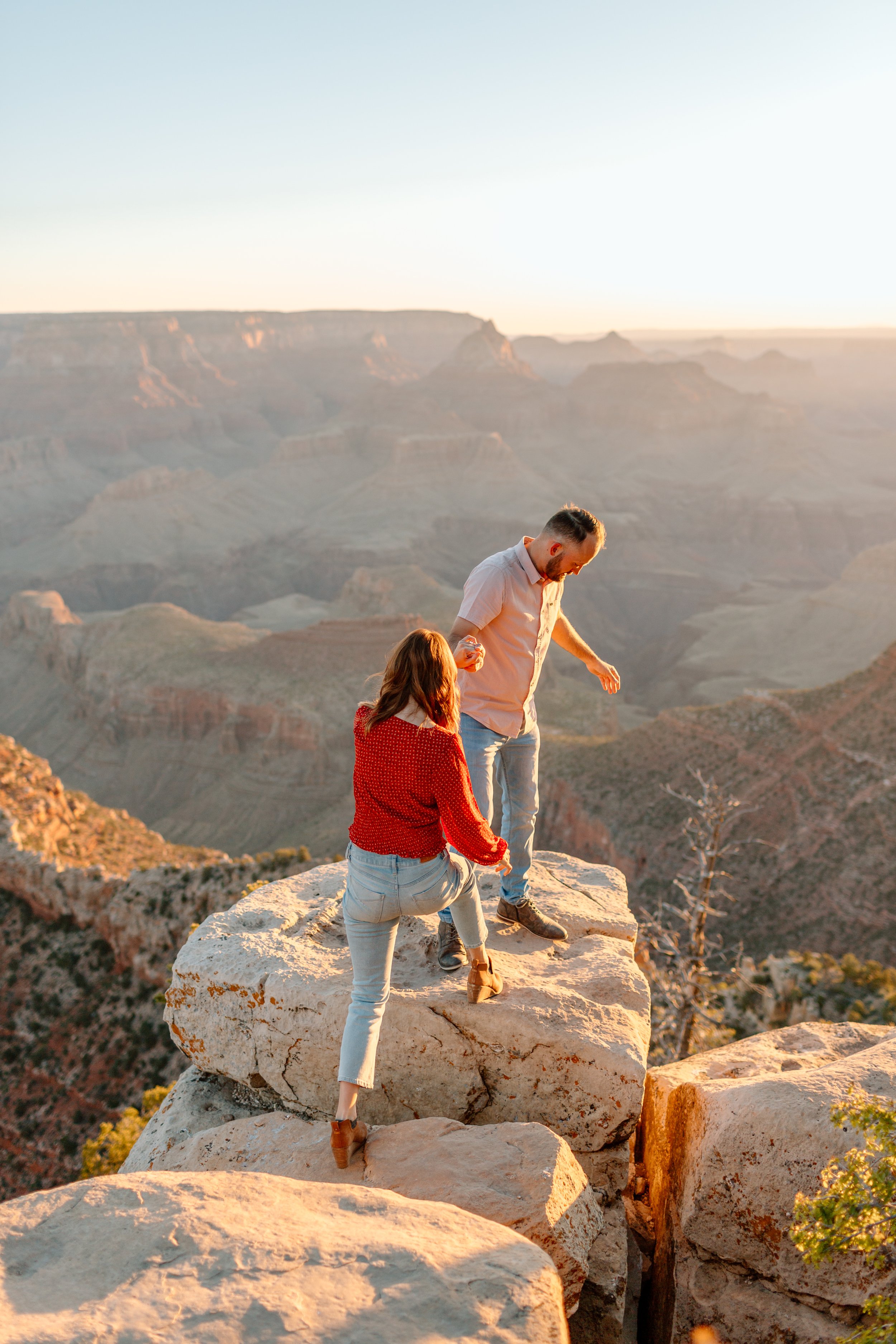  man helps woman up onto a rock during their grand canyon engagement session 