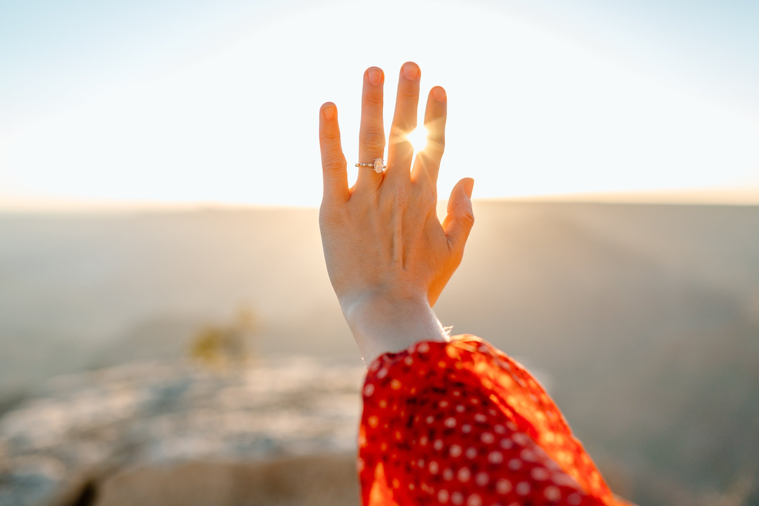  a woman’s hand with her engagement ring and the sunrise peeking between her fingers 