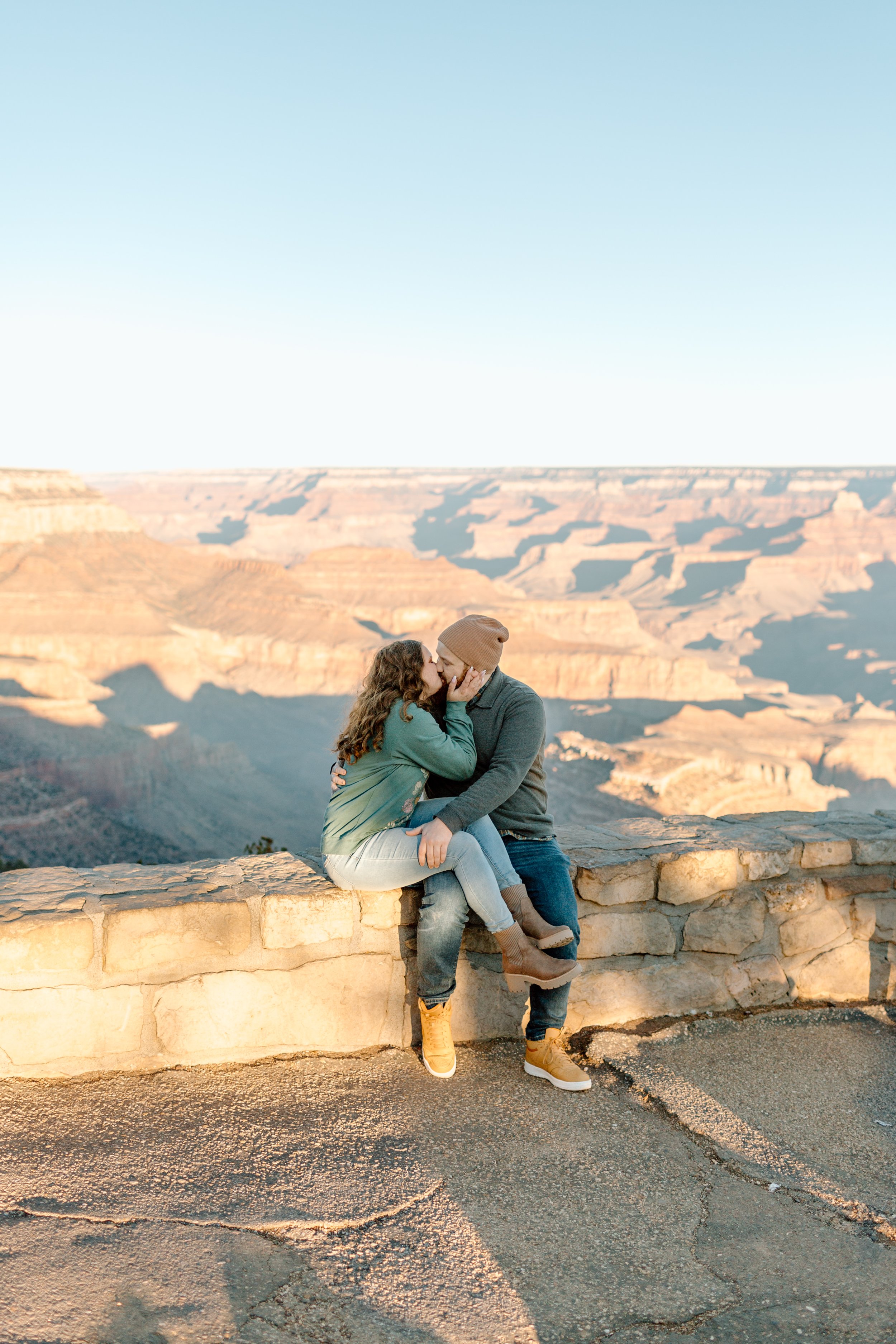  couple sits on a ledge and kisses in front of the grand canyon 