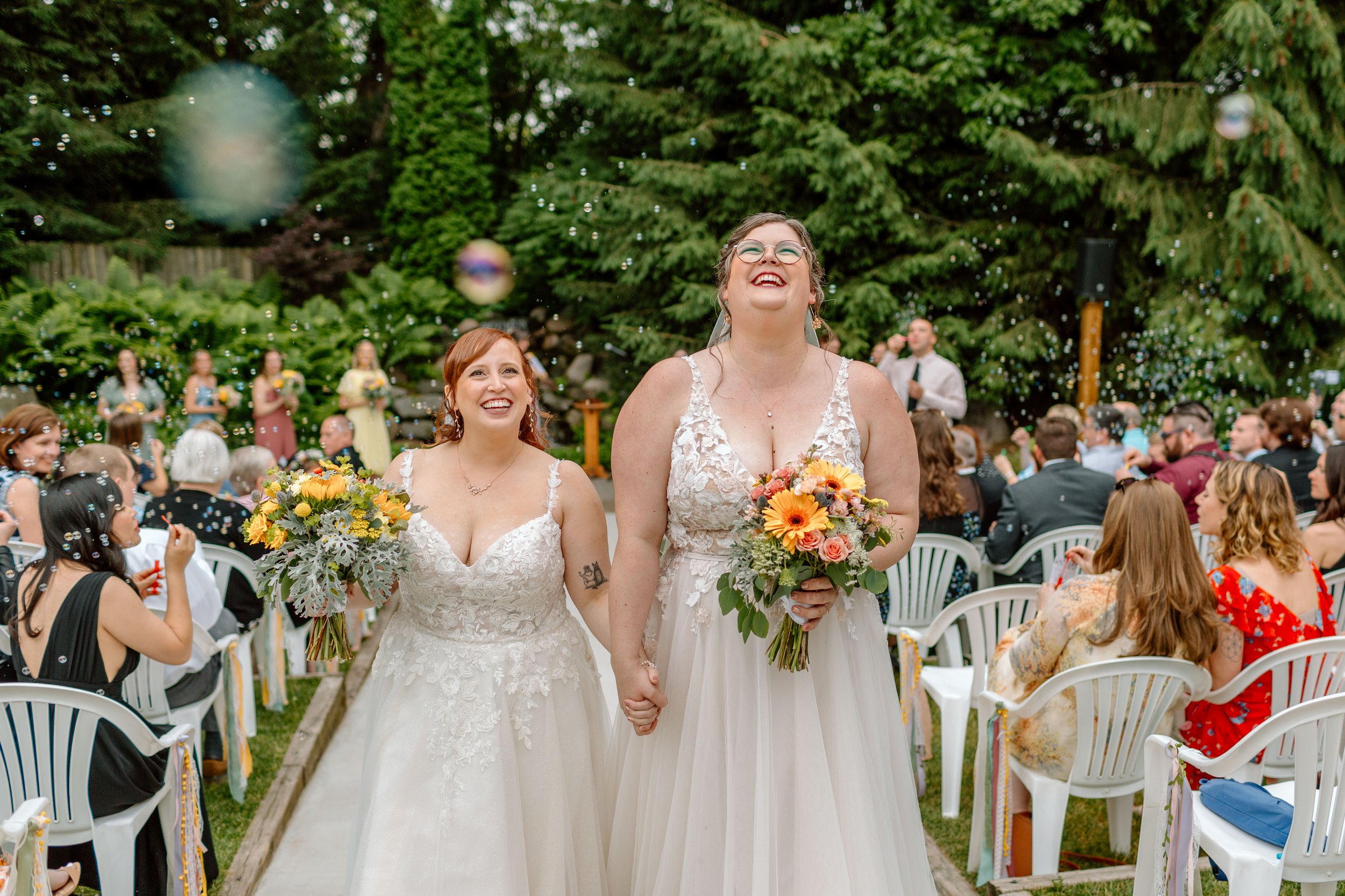  two women walk back up the aisle after getting married, smiling as they are surrounded by bubbles 