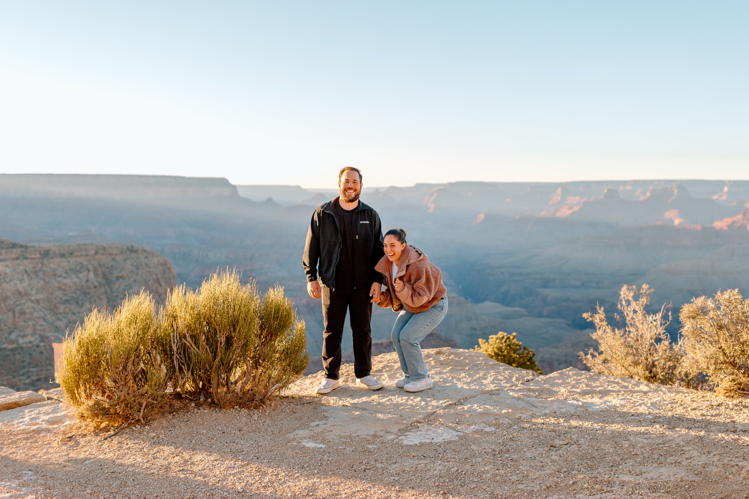  couple stands on south rim of grand canyon at sunset, woman is happily screaming because they just got engaged 