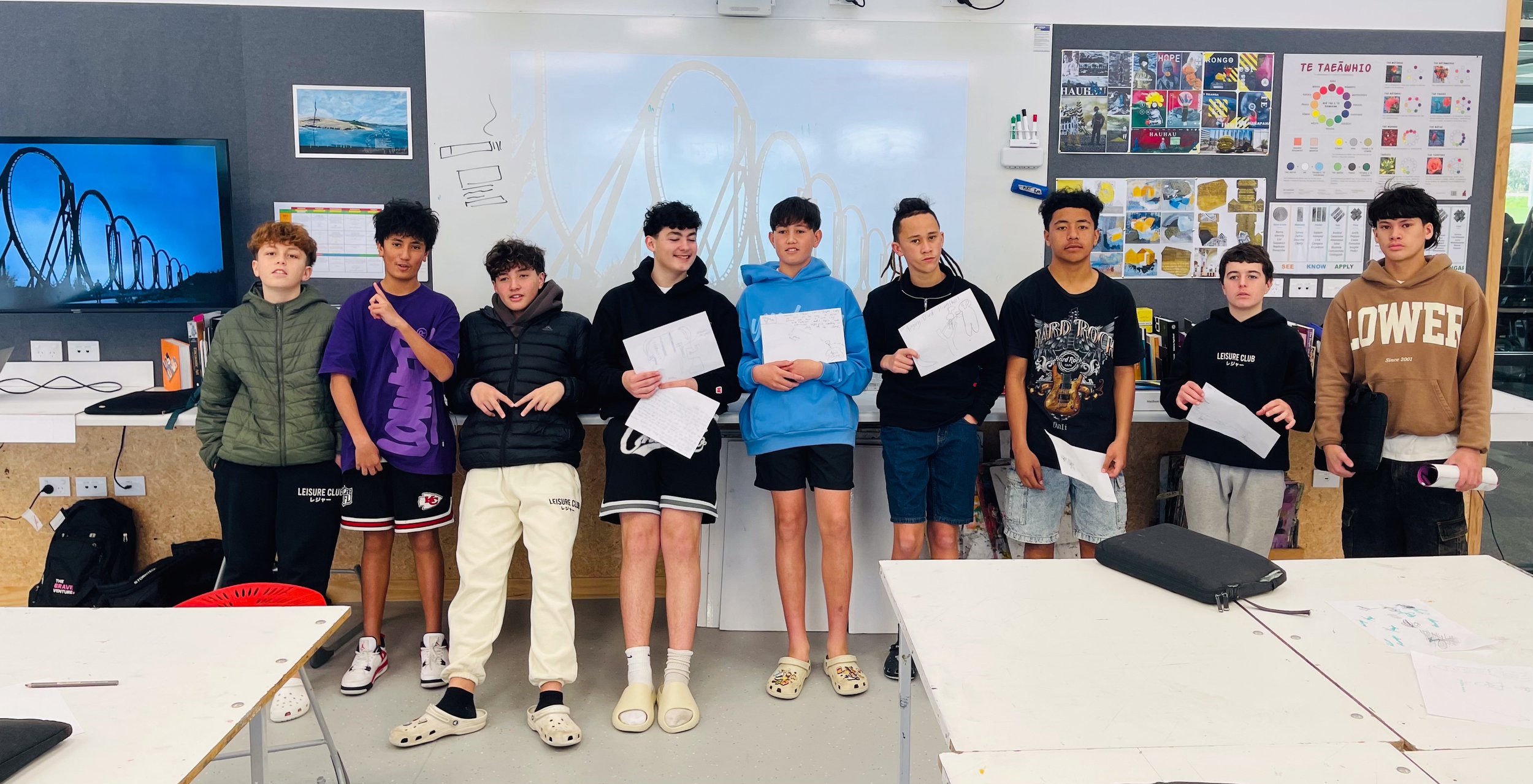 Group of teenagers standing in a classroom holding papers with roller coaster images on the monitor and wall behind them.