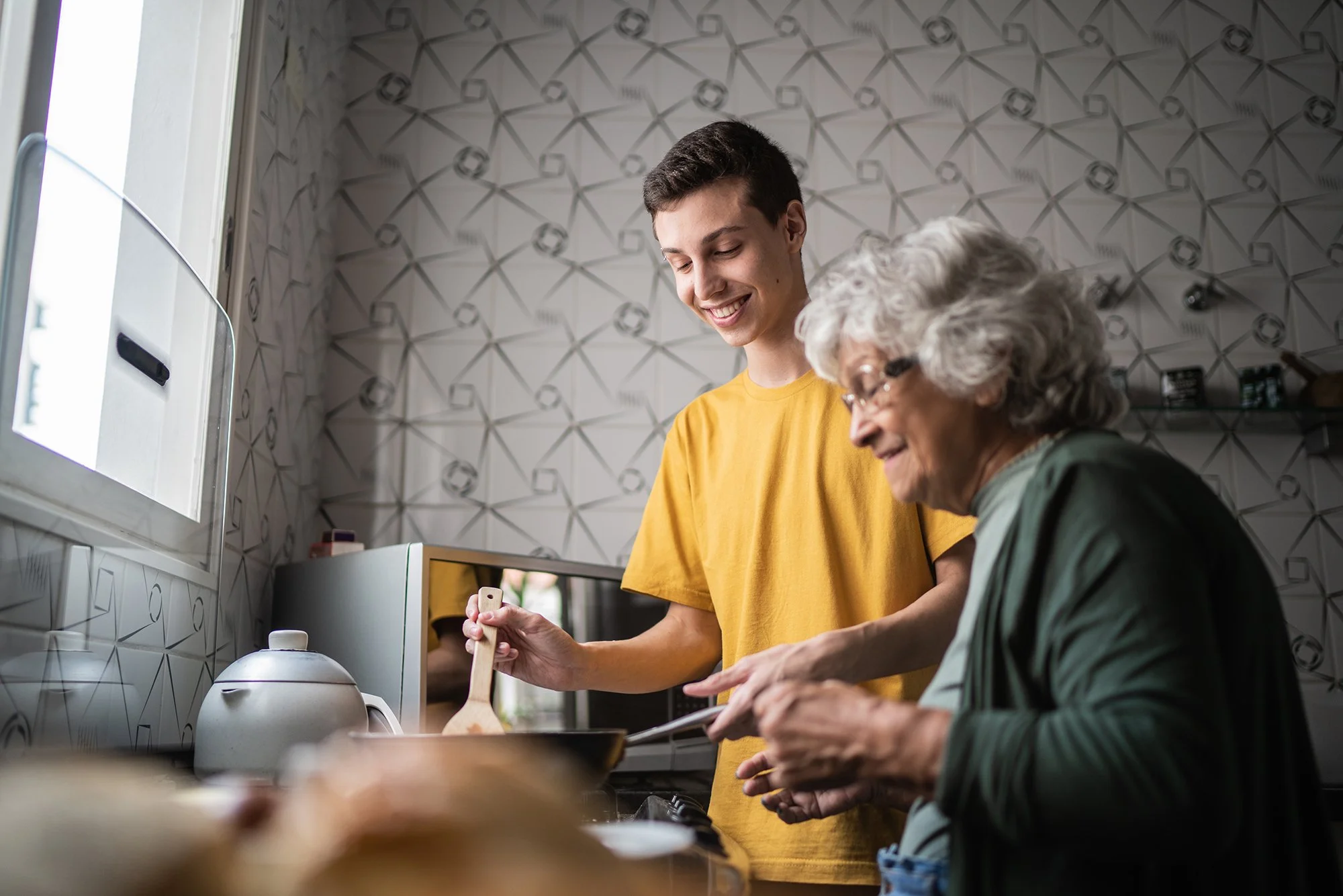 A young man and an elderly woman cooking together in a kitchen, smiling and enjoying their time.