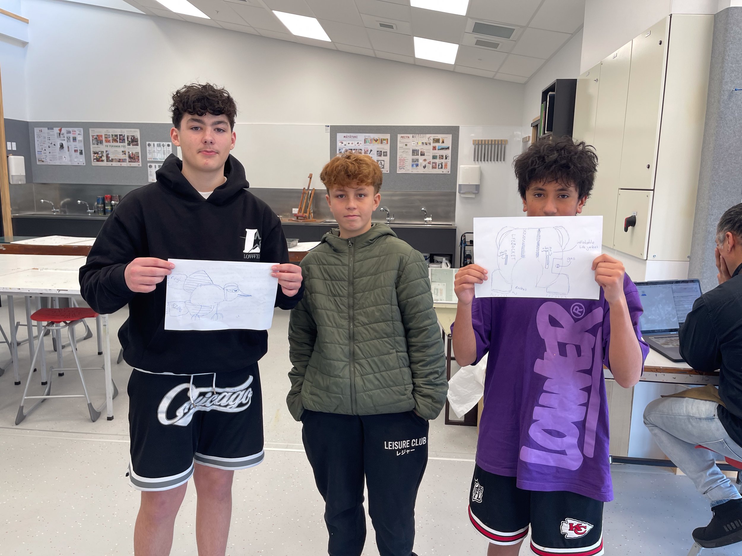 Three adolescents in a classroom, two holding sketches of birds and anatomical drawings, wearing casual attire.