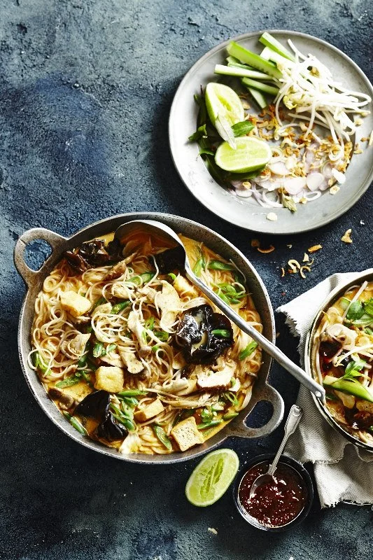 A pot of chicken noodle soup with vegetables and noodles, a bowl of sliced green onions and bean sprouts with lime wedges, and a small bowl of red sauce on a dark surface.