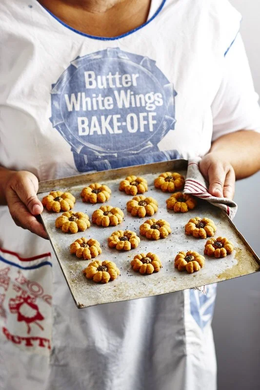 Person in a white t-shirt holding a baking tray with flower-shaped cookies with dark centers. The t-shirt has a blue and white logo that reads 'Butter White Wings BAKE-OFF'.