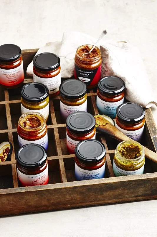 A wooden tray holding multiple jars of Indian spice pastes and sauces with a serving spoon, some open and some closed, on a light surface.