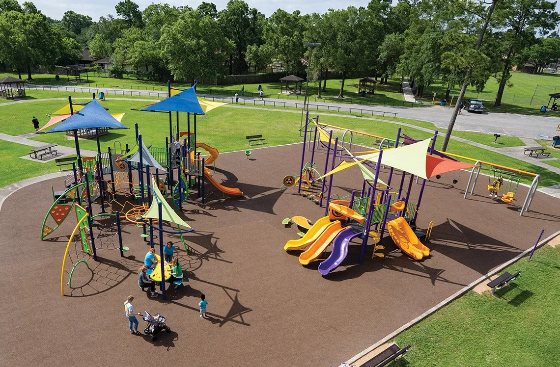 Colorful playground with slides, climbing structures, and shade sails in a park, surrounded by green grass, trees, and picnic tables.