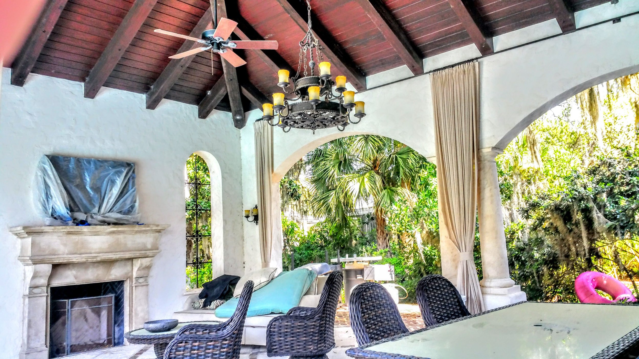Covered outdoor patio with white stucco walls, a dark wooden vaulted ceiling with ceiling fan, and a chandelier with yellow lights. There are large arches, curtains, a fireplace with a stone mantle, wicker chairs around a glass table, and lush greenery outside.