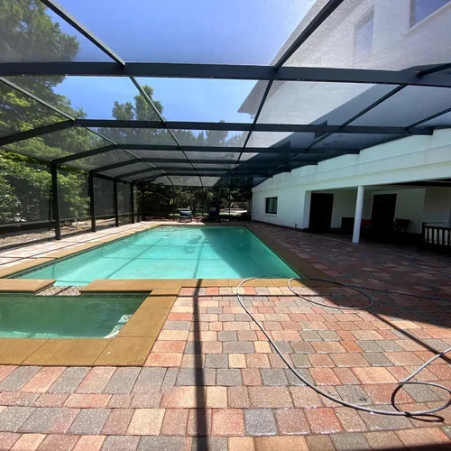 Swimming pool enclosed by a screened-in patio with brick pavers, adjacent to a white house wall with a door and windows, under a clear blue sky.