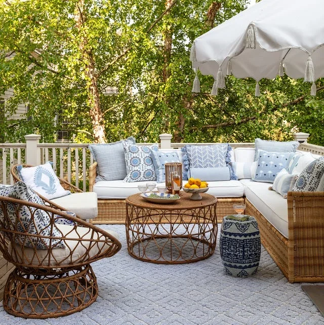 Outdoor patio seating area with a white sectional sofa, wicker chairs, and blue and white cushions. There is a wicker coffee table with a lantern, bowls, and drinks, and a patterned ceramic stool. A large white umbrella shades the area, surrounded by green trees.